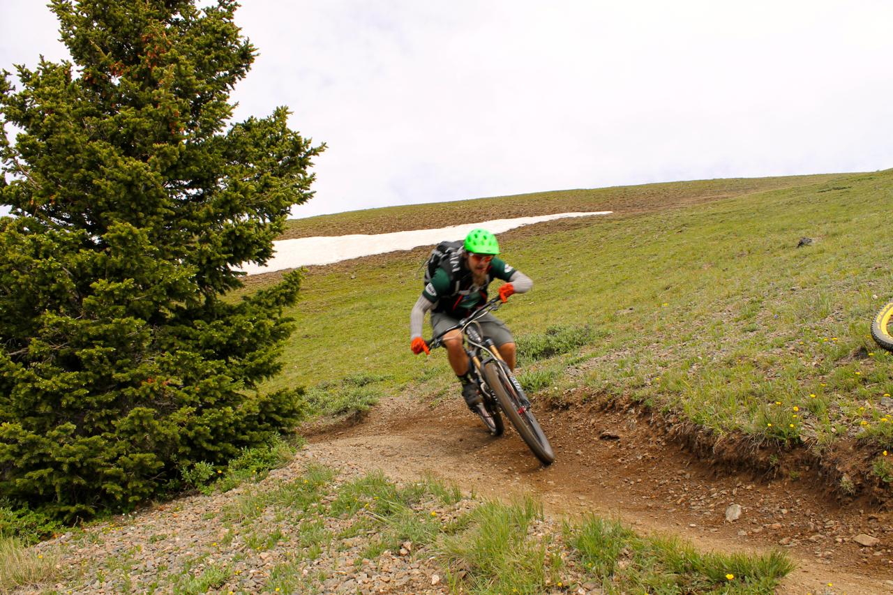 A mountain biker wearing a green helmet and orange gloves navigates a dirt trail surrounded by greenery and patches of snow. Trees are visible in the foreground, and the biker appears to be leaning into a turn, showcasing dynamic movement against a backdrop of a grassy hillside. Monarch Crest Trail mountain bike trail.
