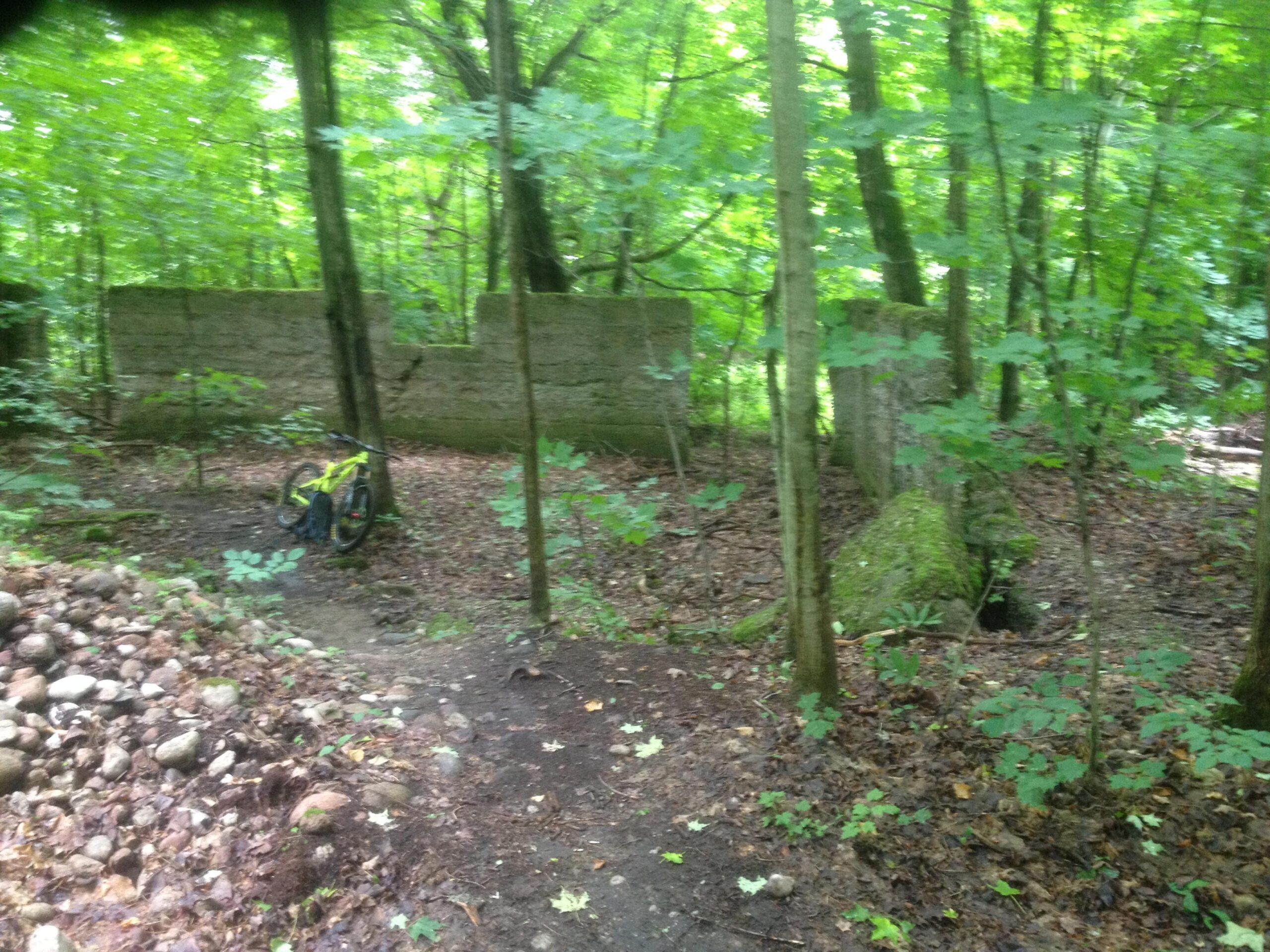 A mountain bike rests on a dirt path surrounded by lush green trees in a wooded area. In the background, a weathered concrete wall is partially visible, covered with moss, amidst a landscape of rocks and fallen leaves. 7thLine to Sugarbush - Green (Nodes D, E, F, G, H, I) mountain bike trail.