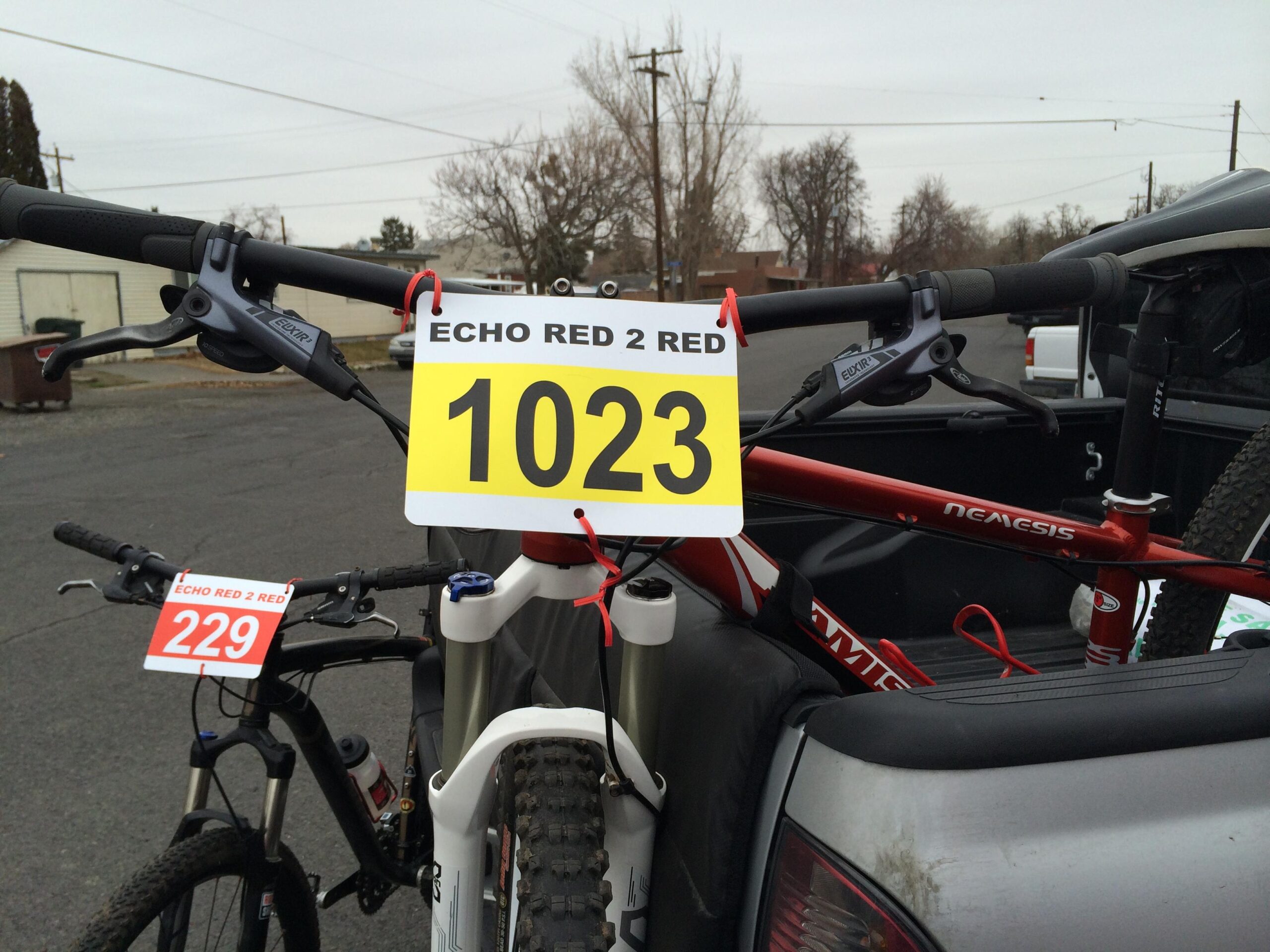 Jamis Nemesis: Alt text: Two bicycles in the back of a pickup truck, each with race numbers attached. The red bike has a number 1023 displayed, while the black bike has a number 229. In the background, there are trees and a cloudy sky.