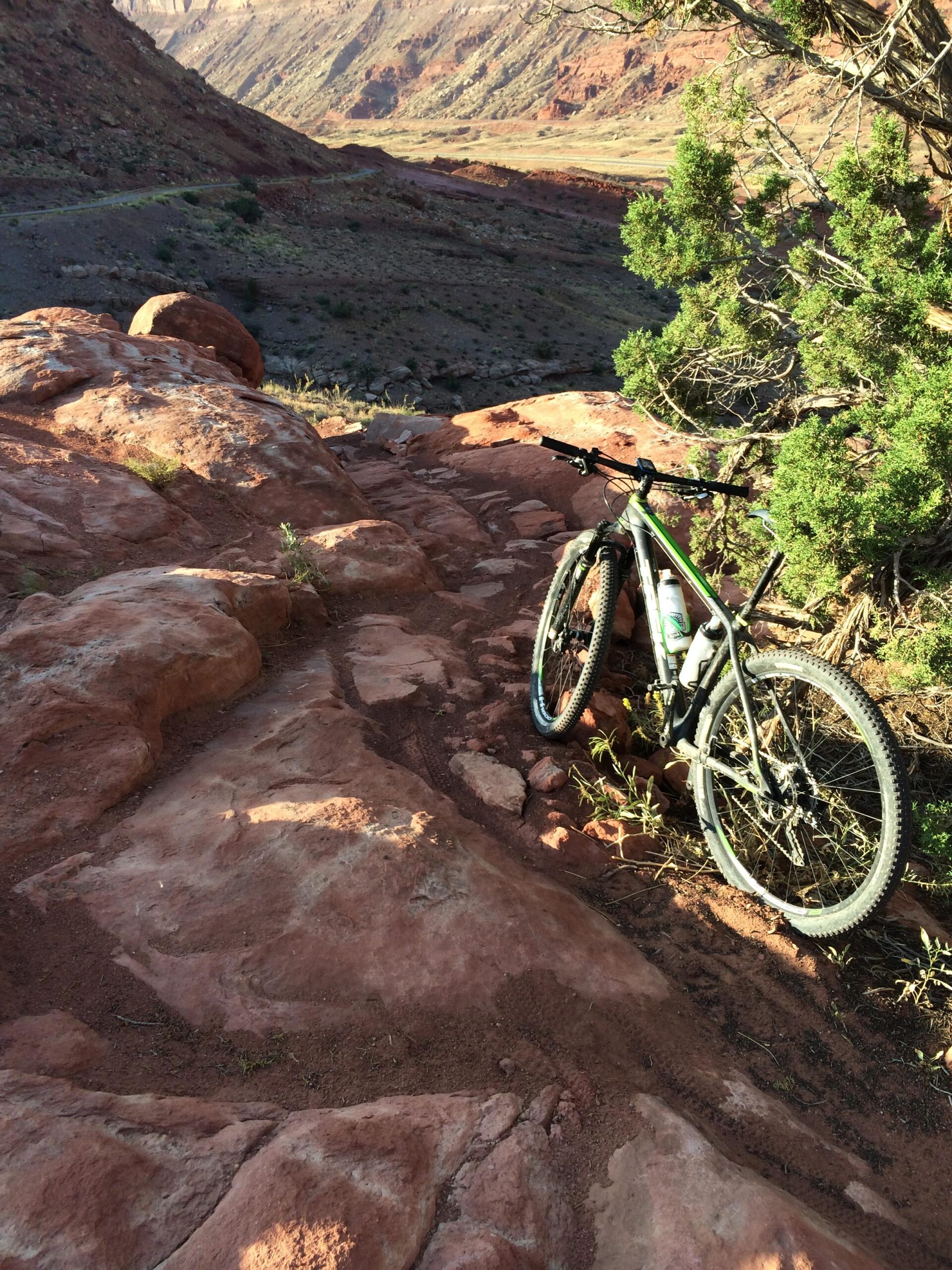 A mountain bike resting on a rocky, uneven trail with a scenic valley in the background, surrounded by reddish-brown hills and greenery. Moab Brand Trails mountain bike trail.