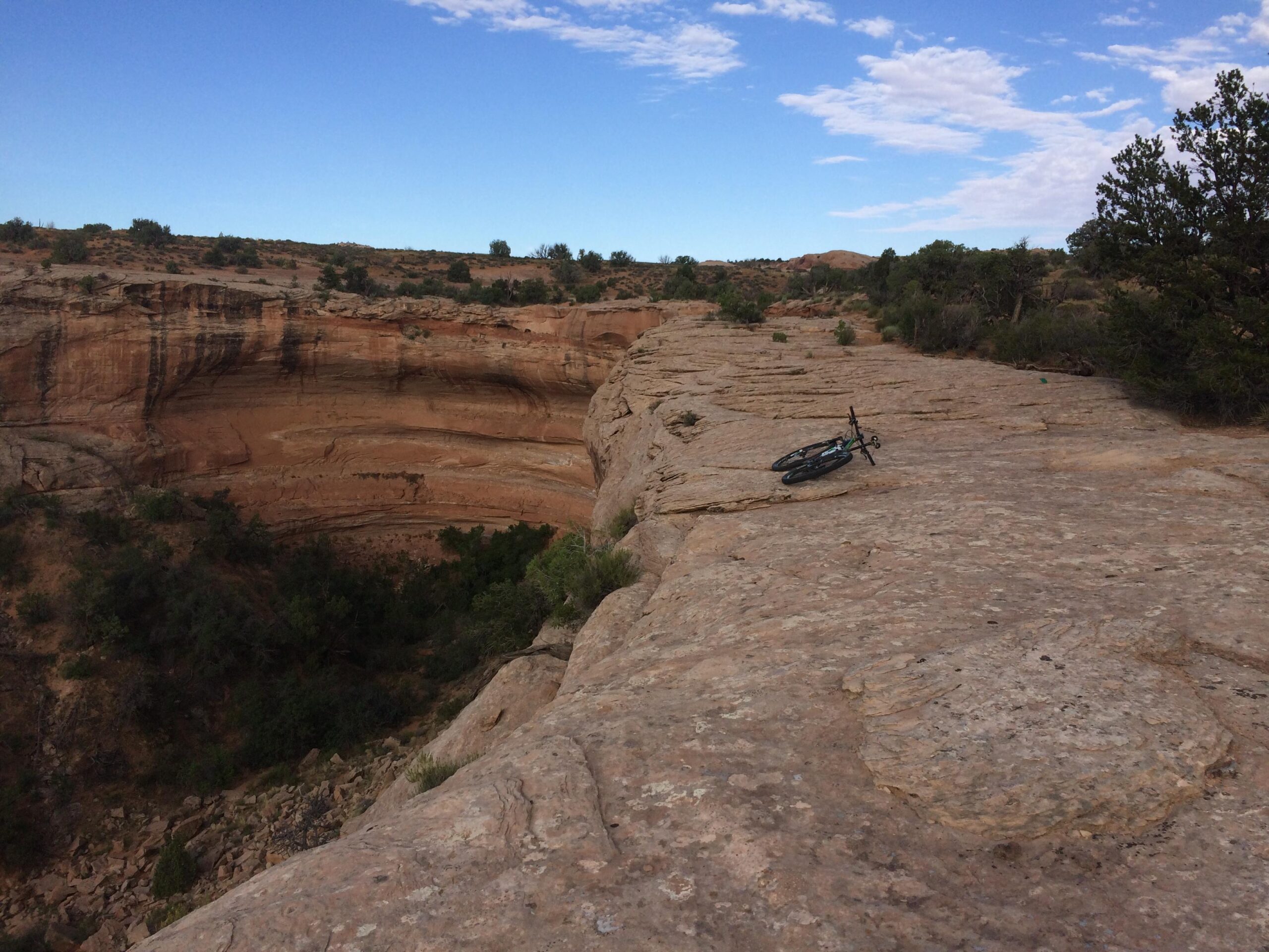 A rocky, rugged landscape featuring a steep cliff edge and a distant canyon below. In the foreground, a bicycle rests on the rocky surface, with sparse vegetation surrounding the area under a clear blue sky with scattered clouds. Navajo Rocks mountain bike trail.