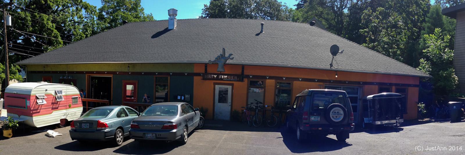 A colorful building with a sloped roof, featuring a sign that reads "Dirty Finger." In front, there are several parked cars and a vintage-style camper trailer. The scene is bright and sunny, with trees and greenery visible in the background.