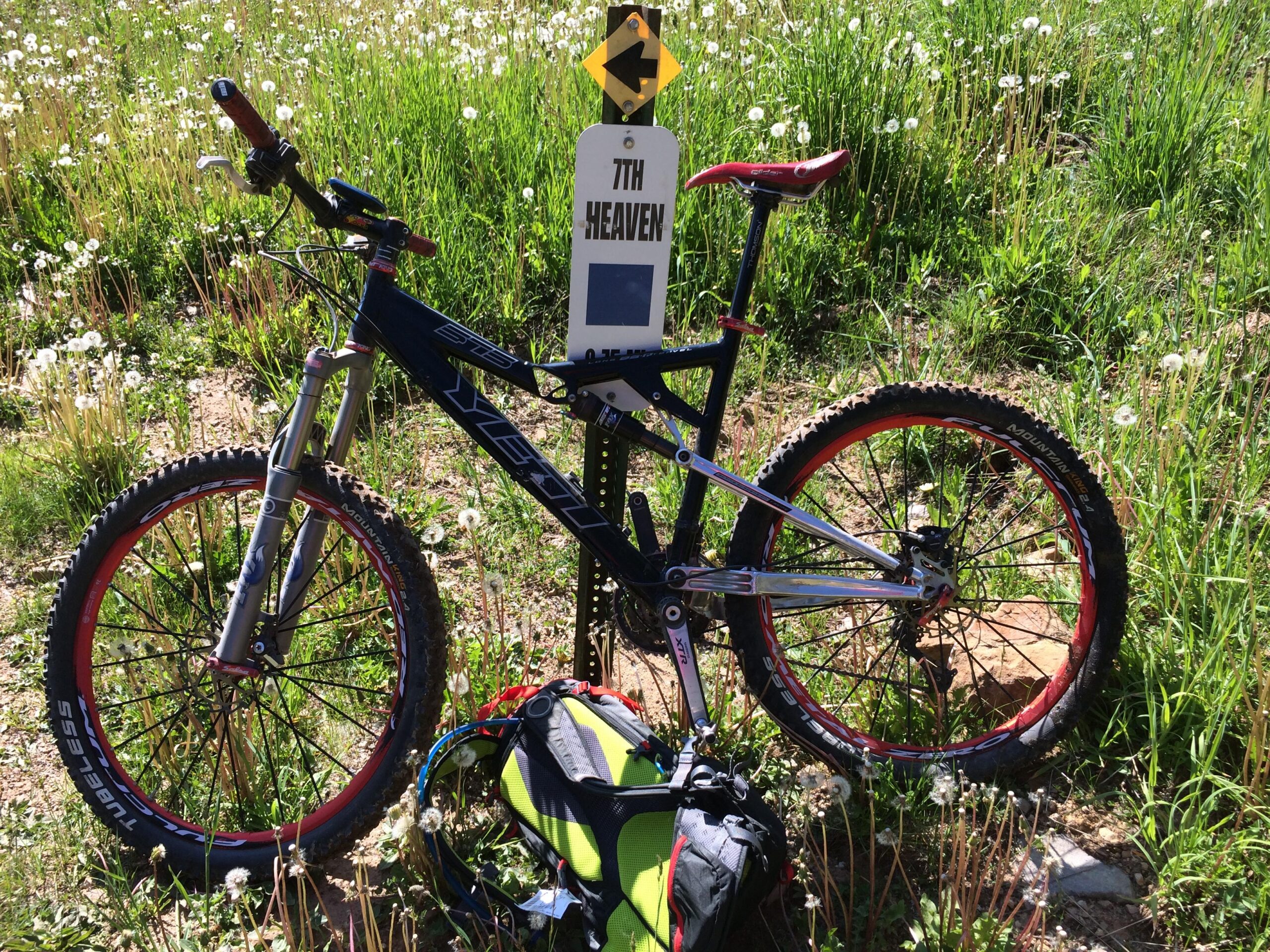 A mountain bike leaning against a trail sign that reads "7th Heaven" in a grassy area filled with dandelions and wildflowers. The bike features red accents on its wheels and handlebars, and a backpack is positioned next to it. 7th Heaven mountain bike trail.