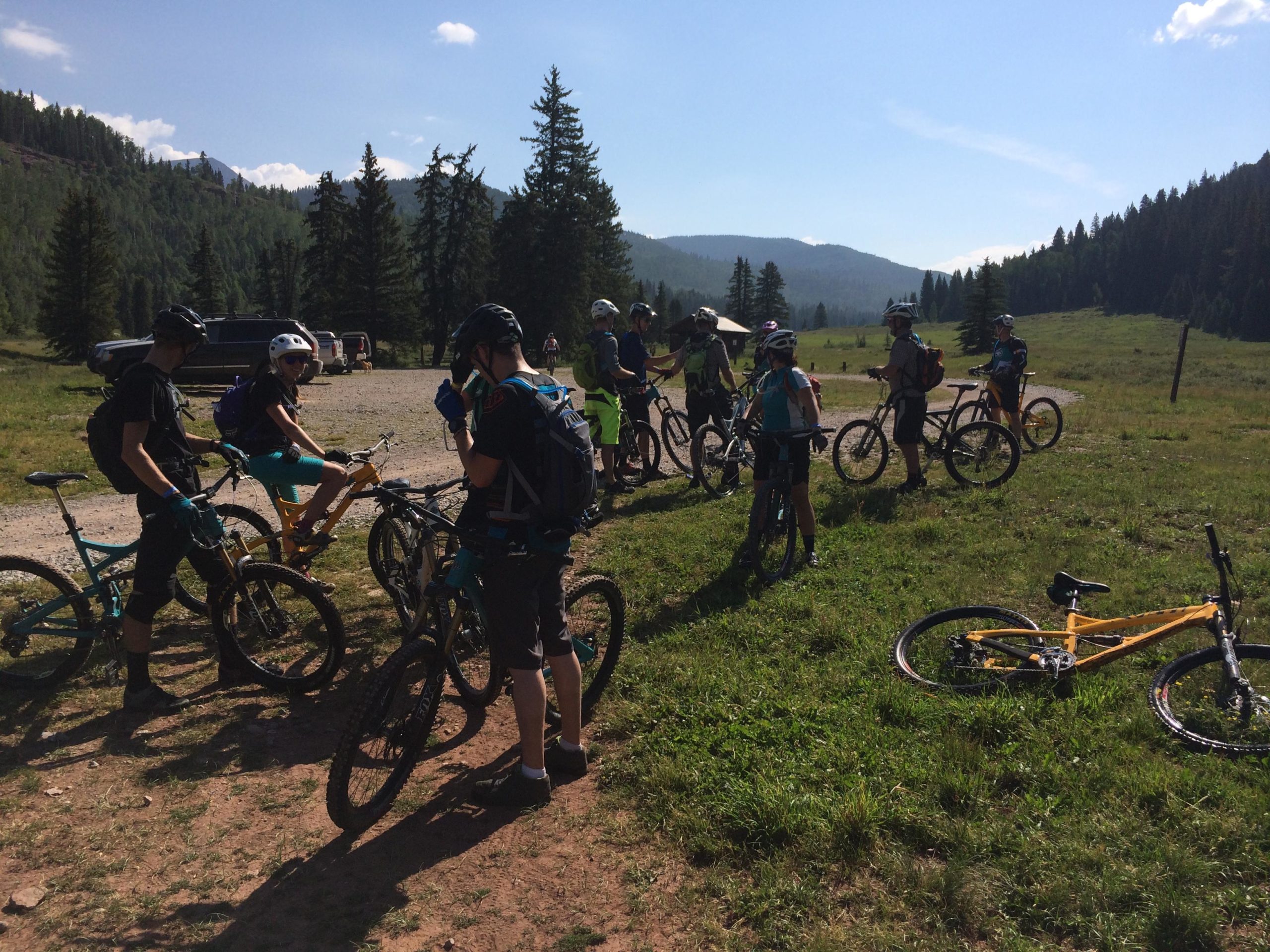 A group of mountain bikers pause for a break in a grassy area surrounded by trees and mountains. Some are standing near their bikes, while one person is seated on a yellow bicycle. The scene is brightly lit with blue skies and a distant view of forested hills. Hermosa Creek Trail mountain bike trail.