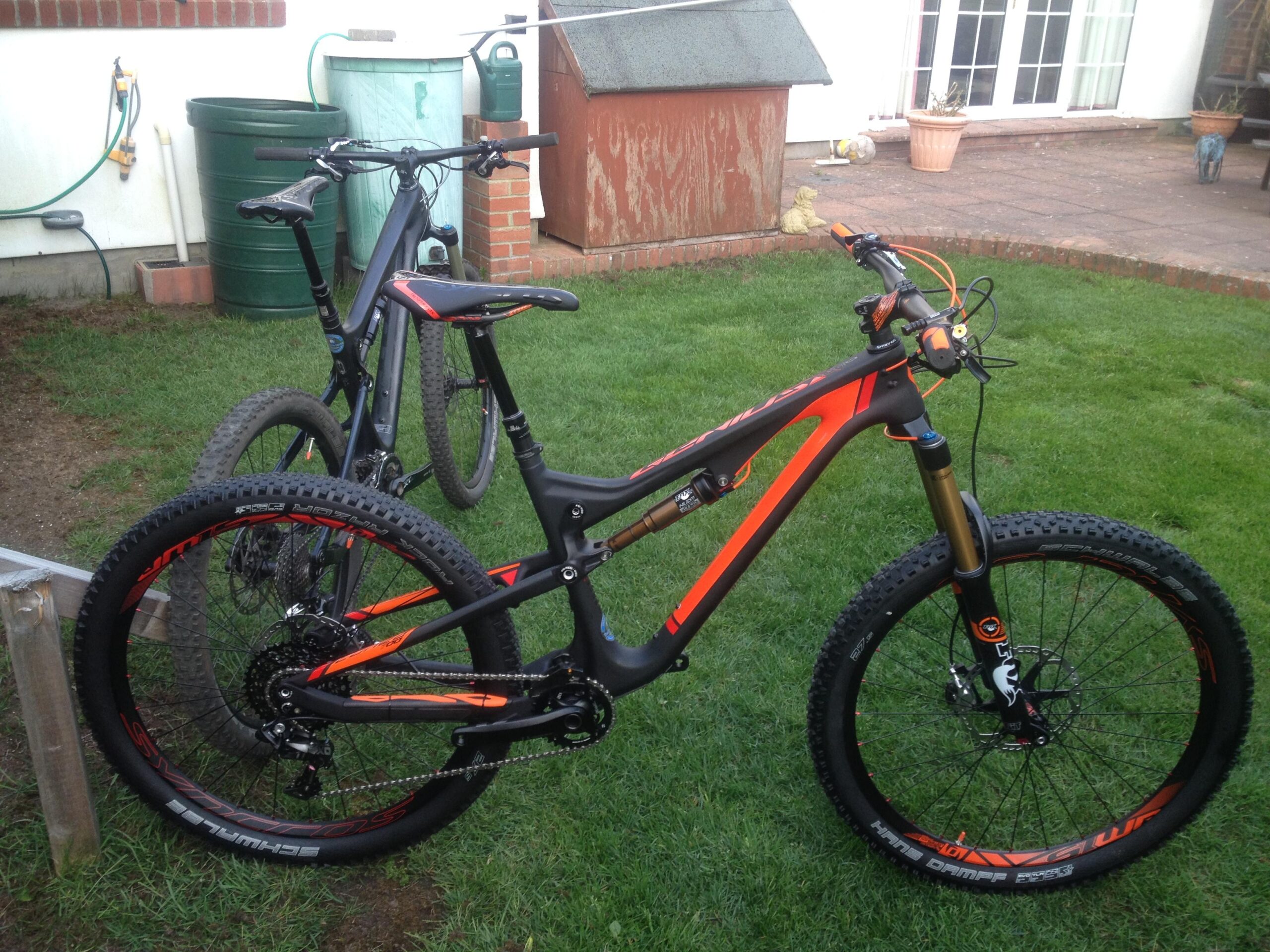 Scott Genius LT 700: A black and orange mountain bike with large tires, positioned on green grass, accompanied by another bike in the background. In the backdrop, there are garden elements, including a storage shed and a watering can.