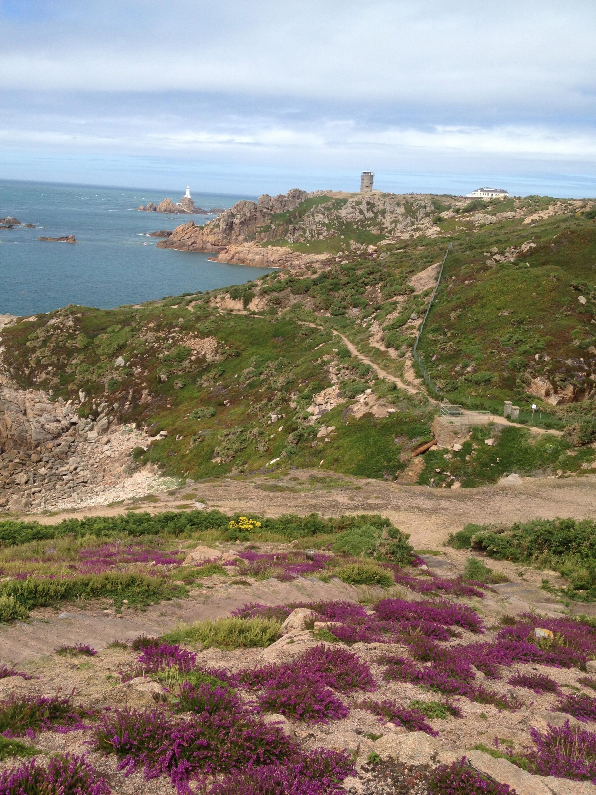A coastal landscape featuring rugged cliffs and green hills dotted with vibrant purple heather. In the background, a lighthouse stands on a rocky outcrop surrounded by the ocean, with a historical tower nearby. The sky is partly cloudy, creating a serene atmosphere. St Brelade loop smash mountain bike trail.