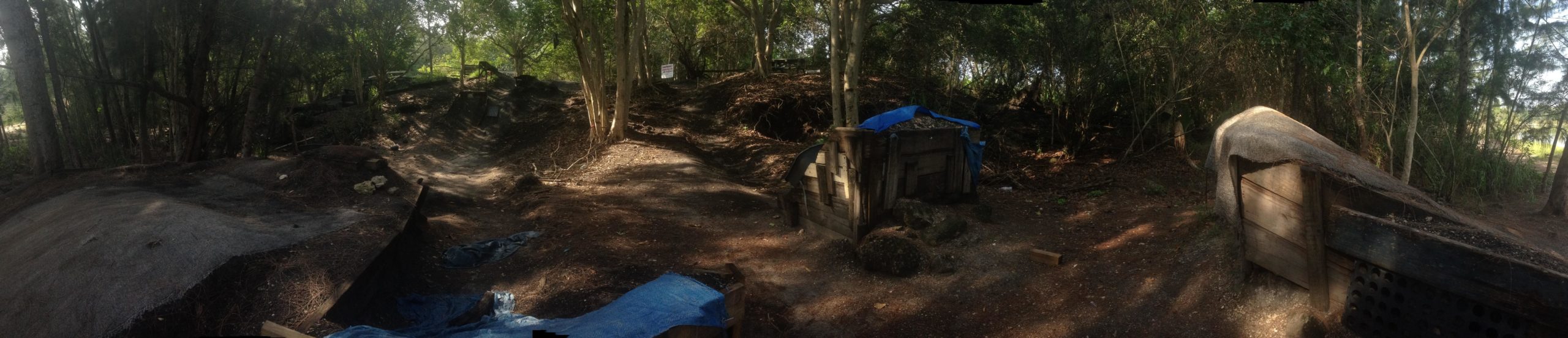 Panoramic view of a wooded area containing makeshift shelters constructed from wood and tarps. The site is surrounded by trees and features uneven ground with patches of dirt, suggesting a rustic or outdoor environment. Sunlight filters through the canopy, creating dappled shadows on the ground. Markham Park mountain bike trail.