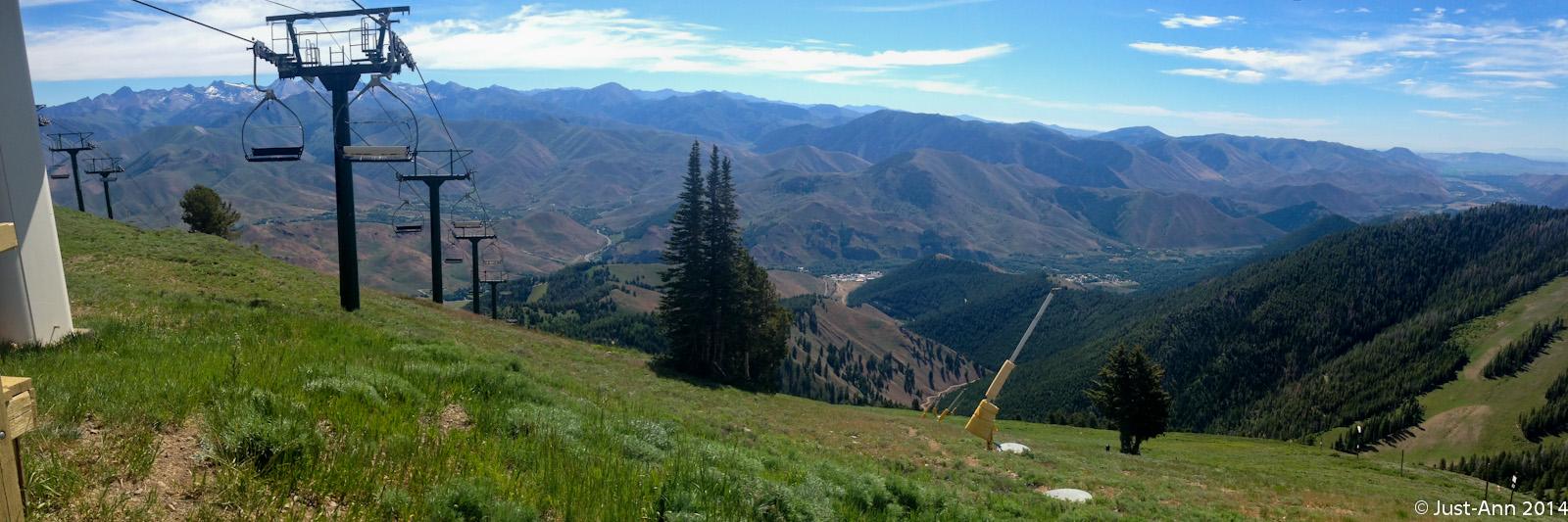 A panoramic view of a mountainous landscape featuring ski lift chairs suspended above green hills and valleys. In the background, snow-capped peaks rise under a blue sky with sparse clouds. The foreground shows lush grass and trees, while the valley below reveals a mix of natural scenery and developed areas. Bald Mountain Bike Park mountain bike trail.