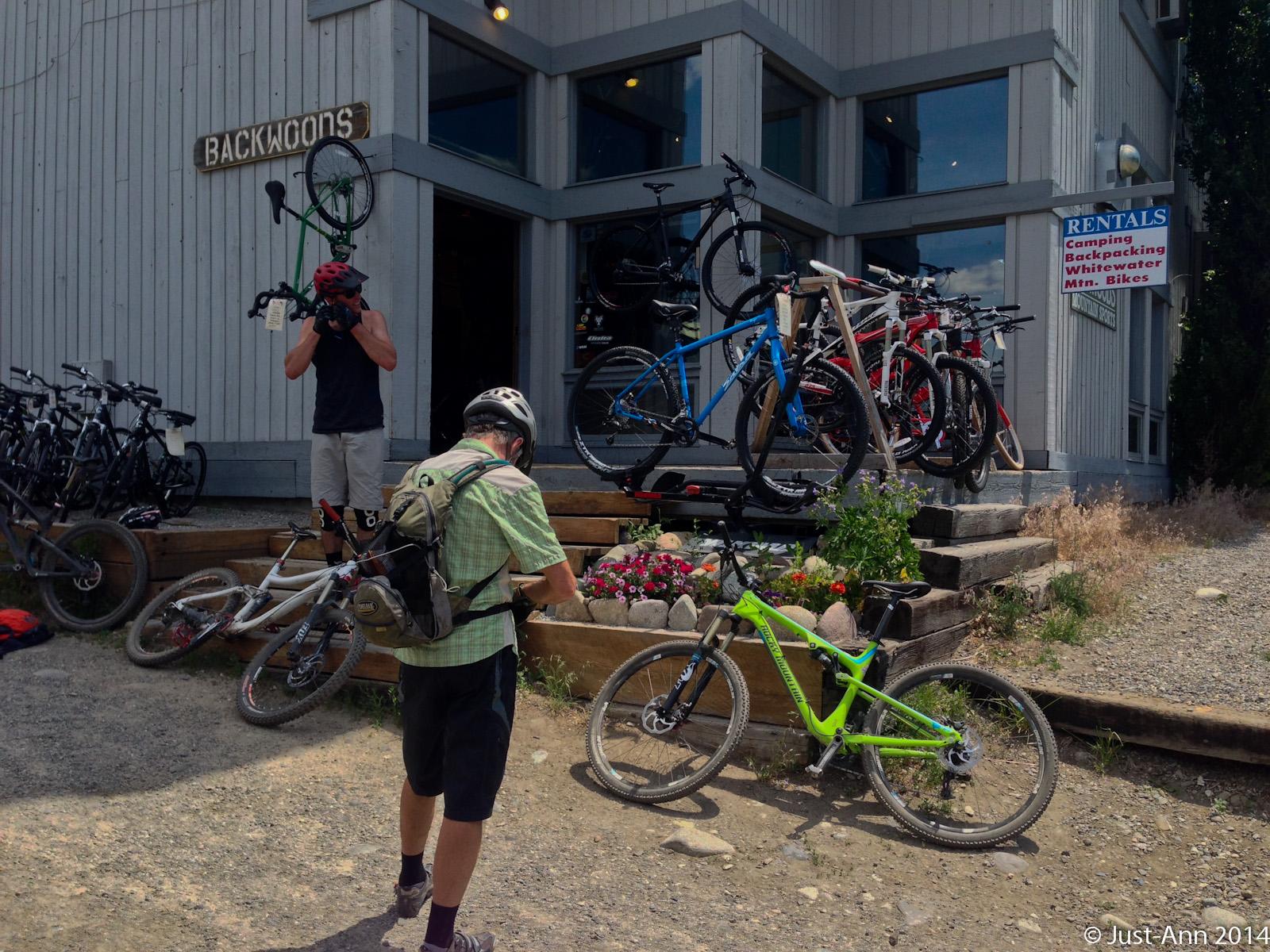 A bike rental shop, Backwoons, featuring a variety of bicycles displayed outside. One person is taking a photo of a bike, while another individual is checking their equipment. The scene includes colorful mountain bikes and flower arrangements in the foreground, set against a backdrop of a wooden building with large windows.