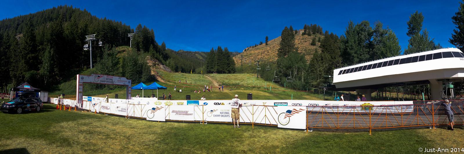 A panoramic view of a mountain biking event setup, featuring a registration area with banners for the National Championships. In the foreground, a grassy area is enclosed by barriers, with a tent and a parked car visible. In the background, a steep hill is dotted with trees and mountain bike riders preparing for the competition. The sky is clear blue, indicating a sunny day. Bald Mountain Bike Park mountain bike trail.