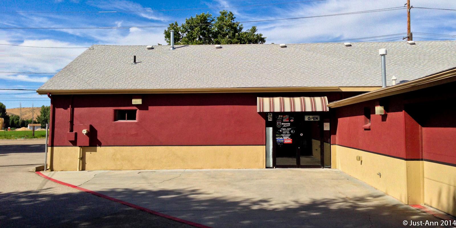 A view of the exterior of a building featuring a red and tan color scheme, with a doorway that has an "Open" sign. The building is partially surrounded by a paved area and has a striped awning above the entrance. In the background, there are trees and rolling hills under a clear blue sky.
