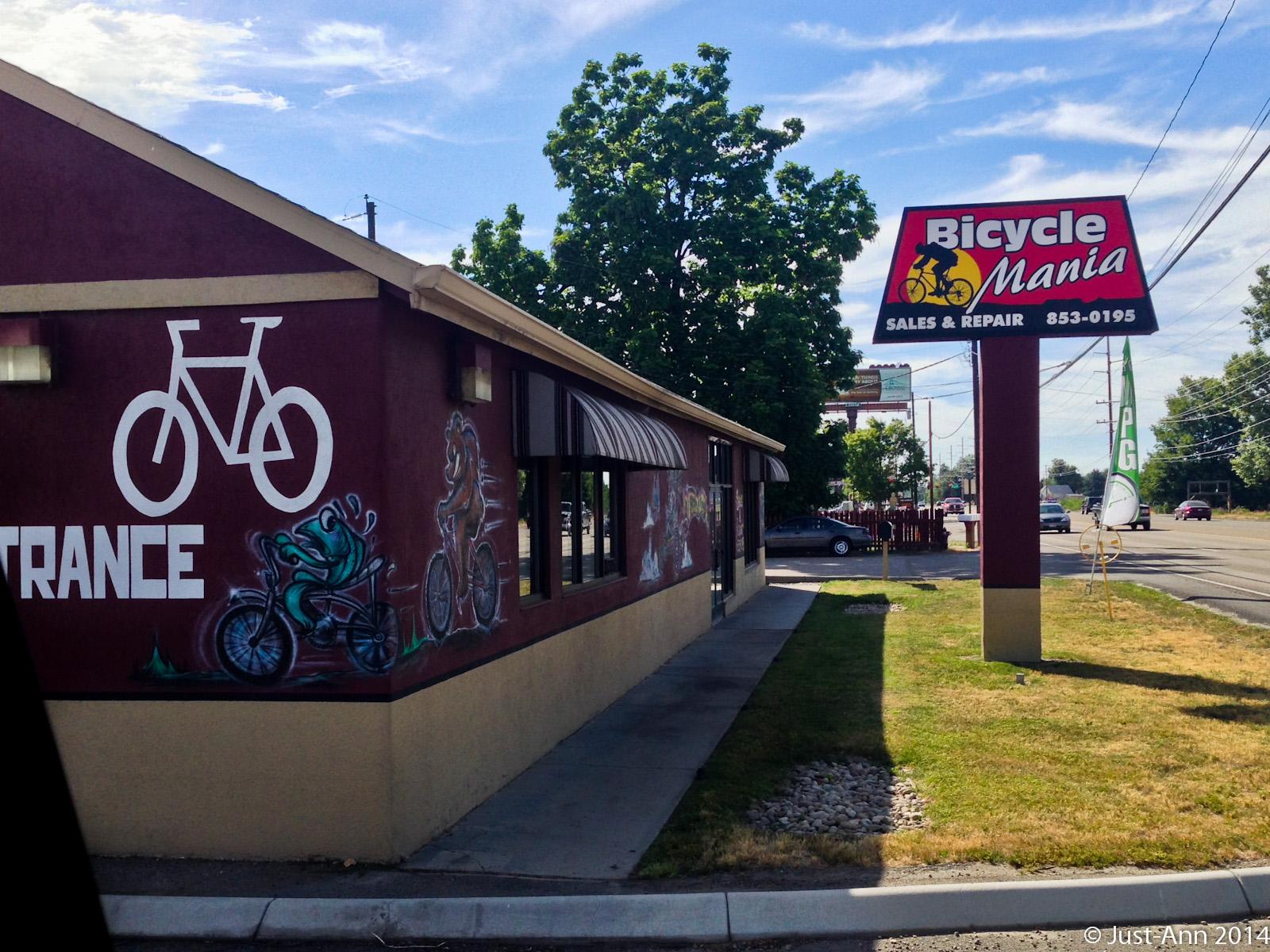 Exterior view of Bicycle Mania, a bicycle sales and repair shop. The building features a large bike graphic and colorful murals on the side. A prominent sign above displays the shop