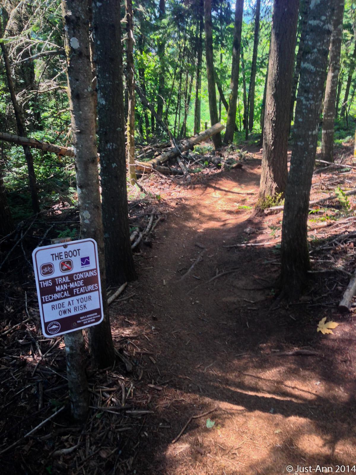 A winding dirt trail through a dense forest, with tall trees on either side. A sign on the left indicates the trail name "The Boot" and warns that it contains man-made technical features, advising users to ride at their own risk. Sunlight filters through the trees, casting shadows on the path. Post Canyon mountain bike trail.