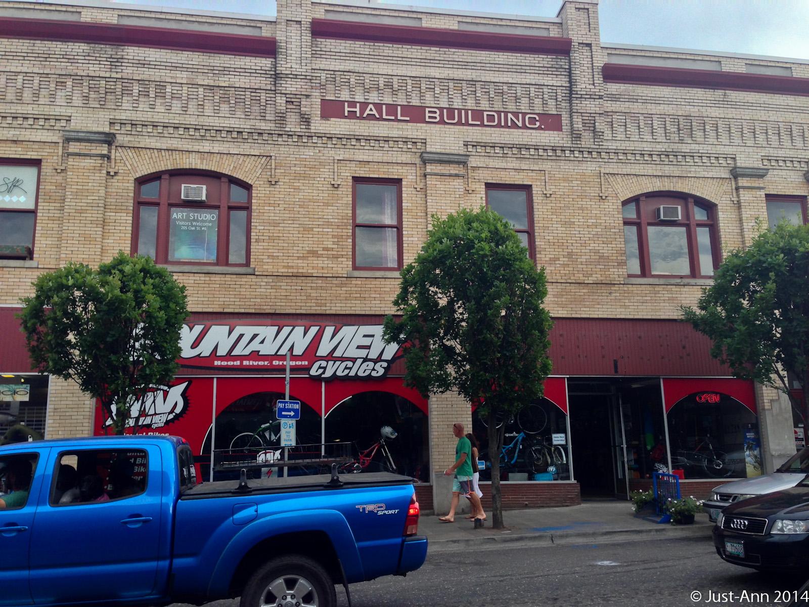 A historic brick building labeled "HALL BUILDING" featuring a sign for "MOUNTAIN VIEW Cycles" on the ground floor. The storefront displays bicycles and has an open sign. There’s a blue pickup truck parked in front, and a person in a green shirt is walking by. The building has large windows and trees flanking the entrance.