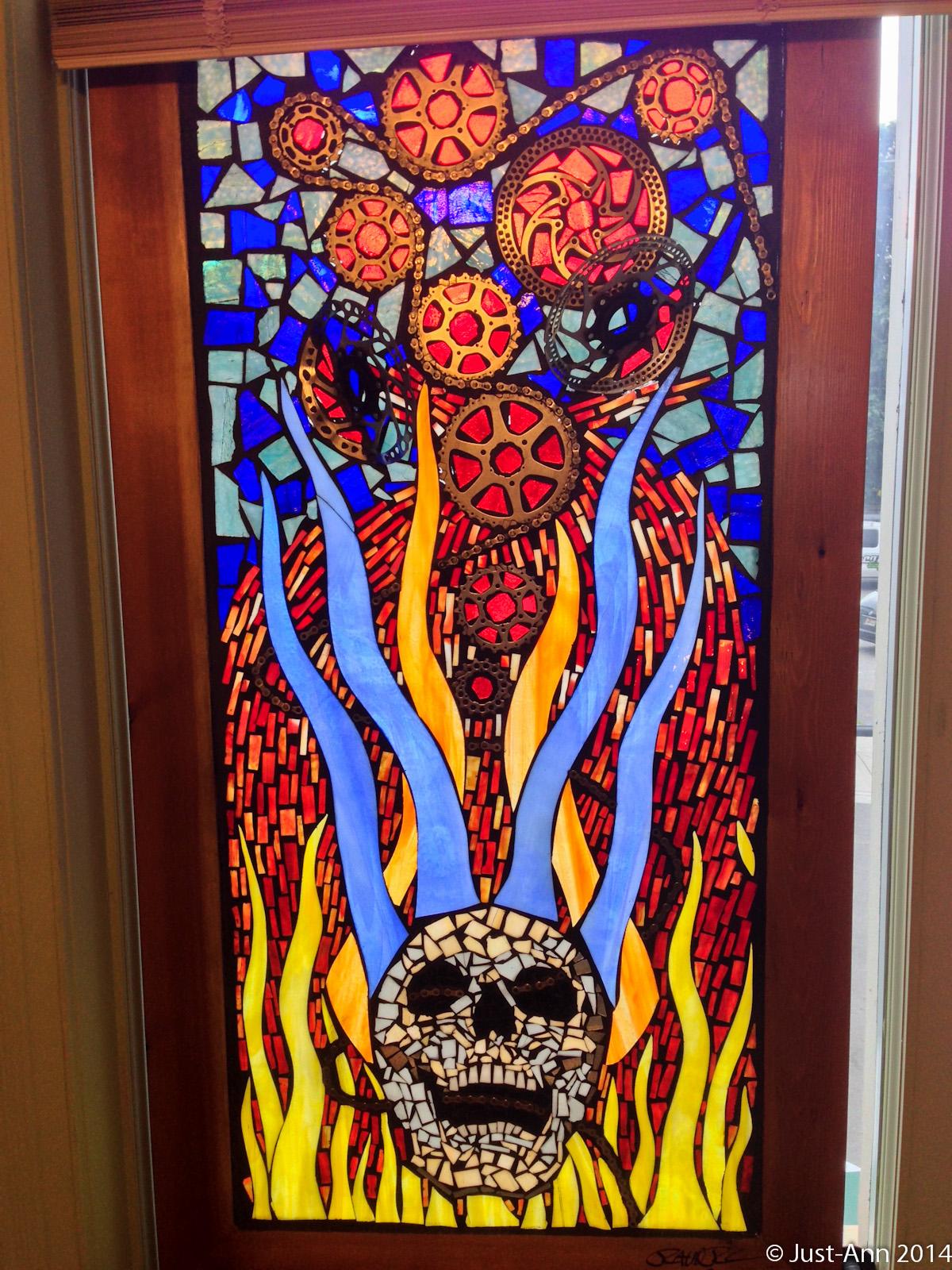 Stained glass window featuring a stylized skull at the center, surrounded by vibrant flames in shades of red and yellow. Above the skull, abstract patterns in blue and orange take the form of circular shapes, set against a mosaic background of blues and blacks. The overall design conveys a striking and dynamic interplay of colors and themes.