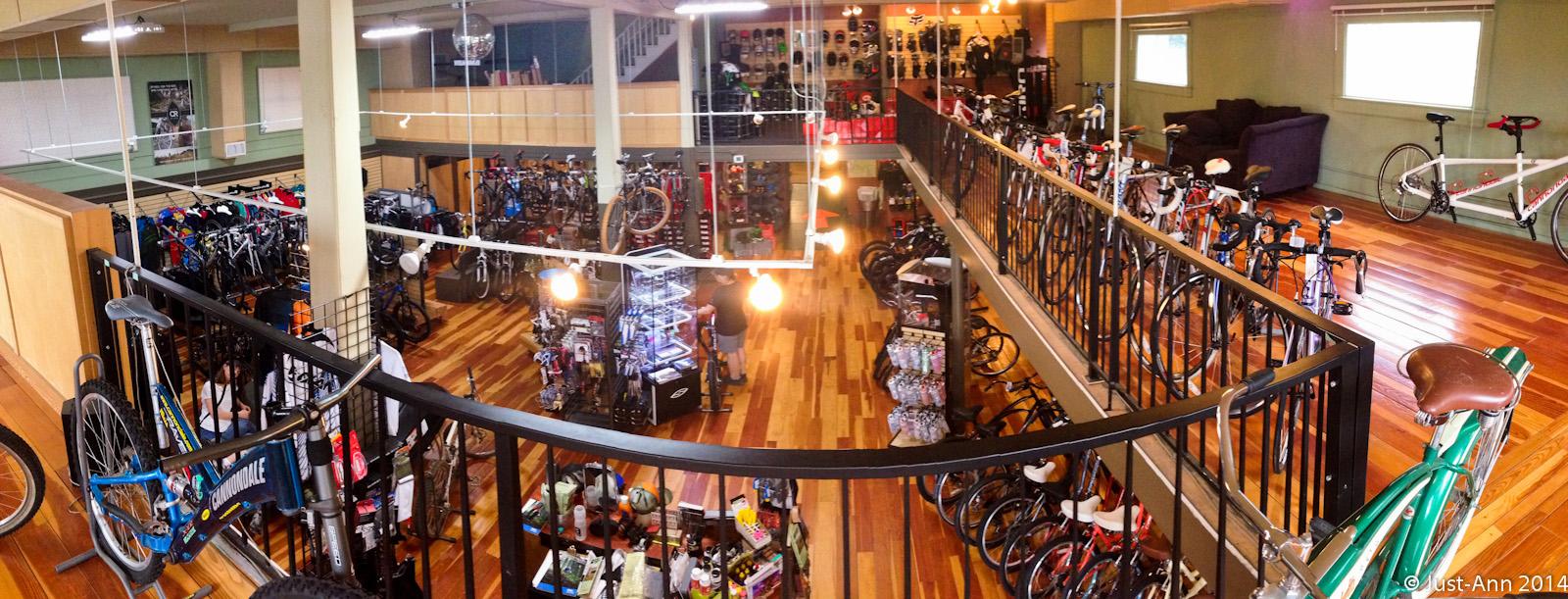 A spacious indoor bicycle shop featuring multiple rows of bikes and cycling accessories. The scene includes wooden flooring, bright lighting, and displays of helmets and gear. A railing overlooks the main floor, where customers are browsing and shopping among a variety of bicycles and equipment.