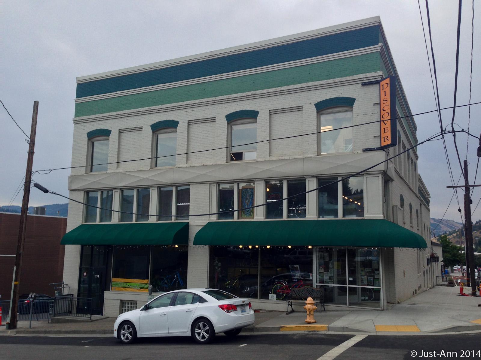 A three-story brick building with green accents and large windows, featuring a green awning over the entrance. A sign that reads "DISCOVER" is prominently displayed on the side. A white car is parked in front, and a fire hydrant is visible on the sidewalk. Power lines run across the image, and a mountain can be seen in the background under a cloudy sky.