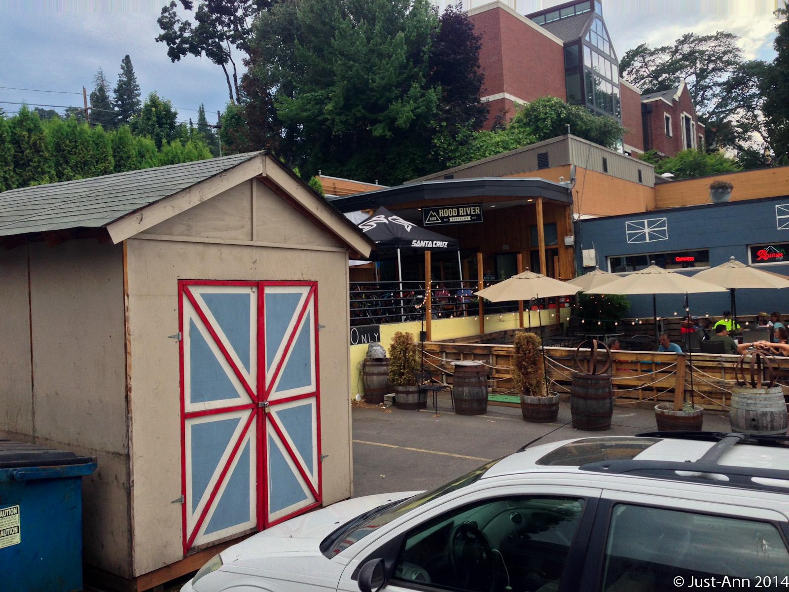 A view of an outdoor dining area at a restaurant featuring a rustic shed painted with a blue and red pattern. The background includes a wooden deck with shaded seating under umbrellas, surrounded by greenery and nearby buildings. A white car is parked in the foreground, alongside a blue dumpster.