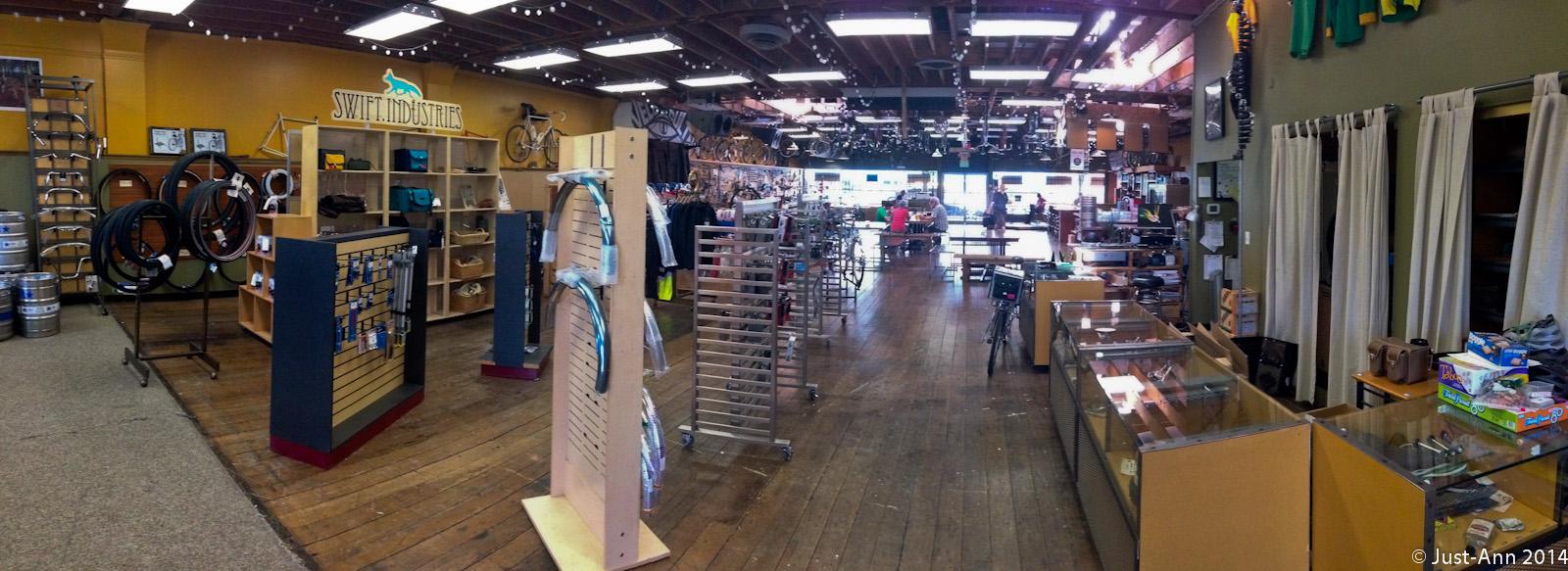A spacious indoor retail store showcasing various biking accessories and gear. The layout features wooden flooring, display racks with bicycle tires, bags, and tools, along with shelves displaying merchandise. Bright lighting highlights the products, and customers can be seen browsing throughout the shop. The store has a modern yet inviting atmosphere, perfect for cycling enthusiasts.