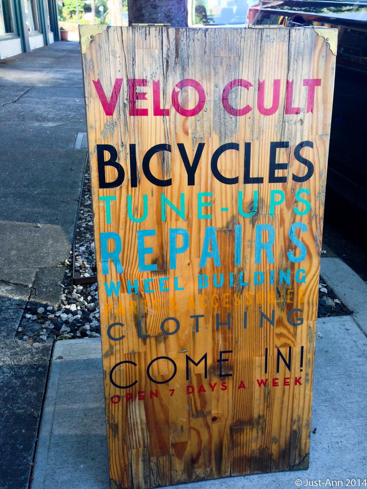 Wooden sign for a bicycle shop featuring colorful text that reads "VELO CULT," "BICYCLES," "TUNE-UPS," "REPAIRS," "WHEEL BUILDING," "PARTS & ACCESSORIES," "CLOTHING," and "COME IN! OPEN 7 DAYS A WEEK." The sign is weathered and stands on a sidewalk in a urban setting.