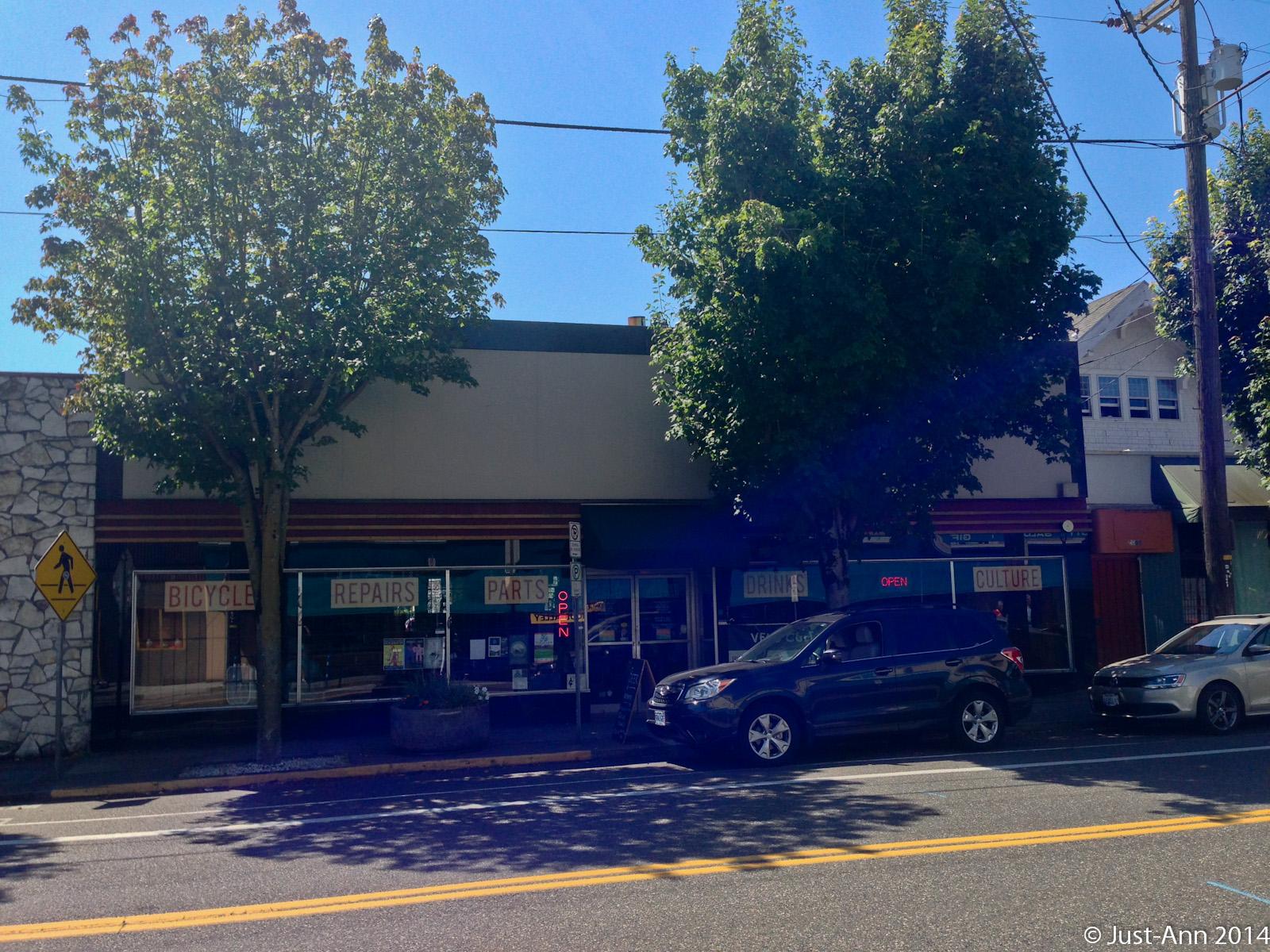 A sunny street scene featuring a row of small businesses. The storefronts include a bicycle repair shop with signs for repairs and parts, and another shop promoting drinks and culture. There are several parked cars along the curb, and green trees line the street under a clear blue sky. A pedestrian crossing sign is visible in the foreground.