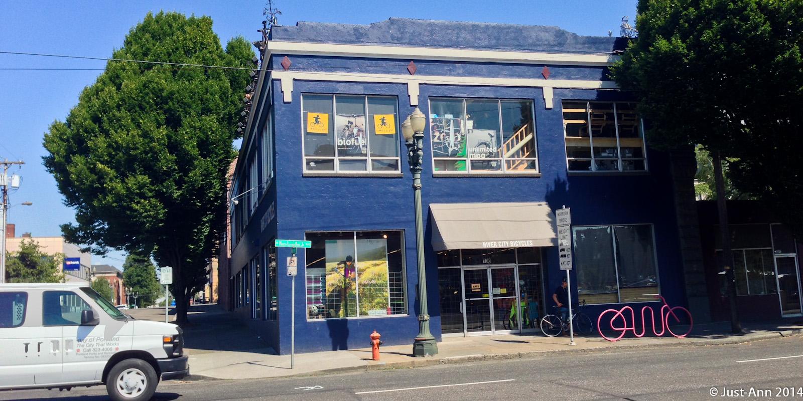 A vibrant blue building featuring large windows displaying bicycle-themed advertisements. In front, there is a unique pink bicycle rack, and a few trees line the sidewalk. A white van is parked nearby, and a cyclist is seen approaching.