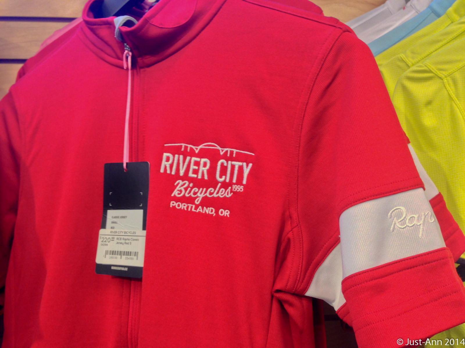A red cycling jersey with the logo "River City Bicycles" and "Portland, OR" embroidered on the front. The jersey features a zippered neck and white accents on the sleeves. A price tag indicating the item is labeled as an "Aero Classic Jersey." In the background, other cycling jerseys in various colors can be seen hanging on a display.