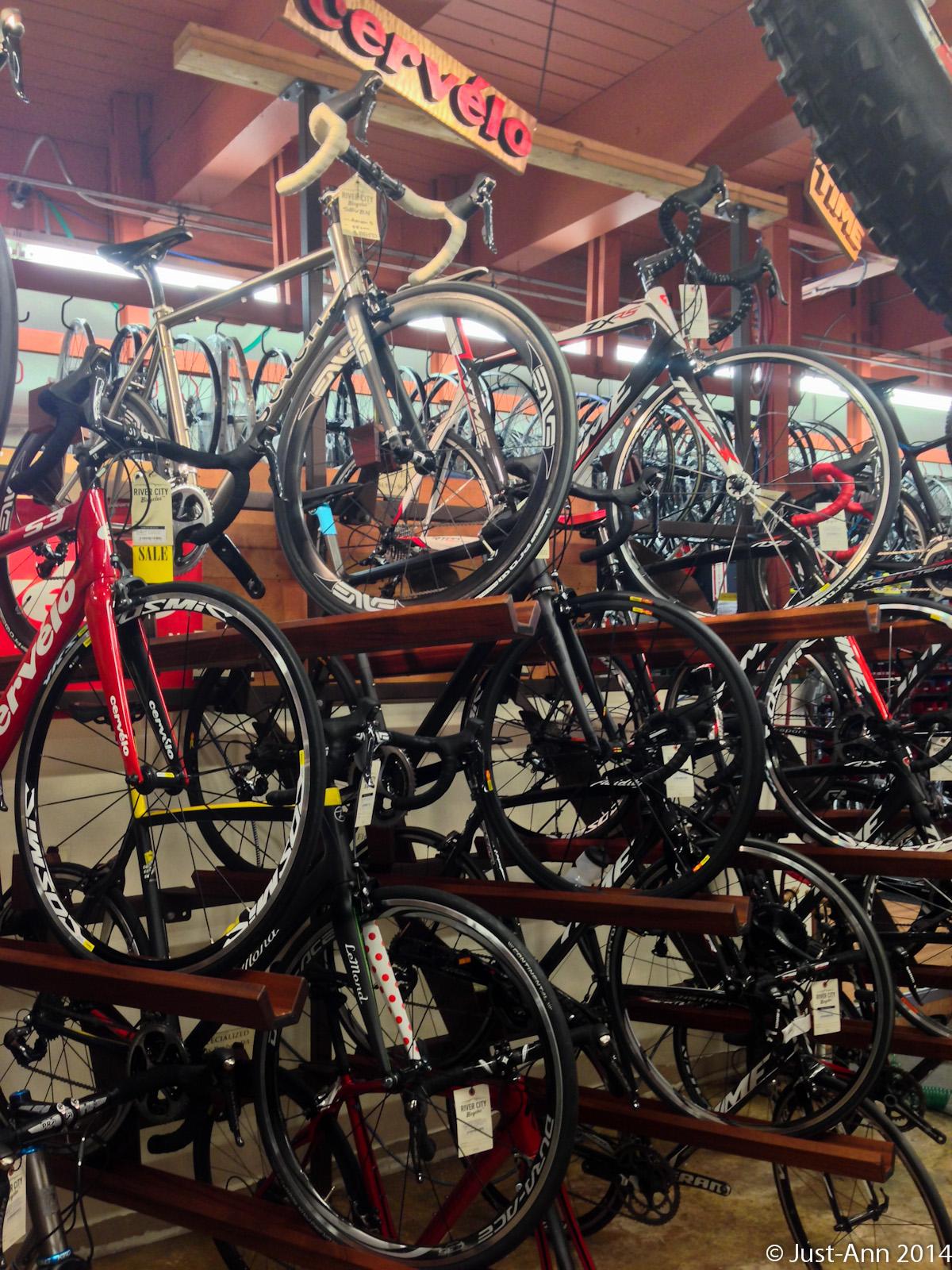 A display of various road bicycles arranged on wooden racks in a bicycle shop, featuring brands like Cervélo and more. The bikes are positioned at different angles, showcasing their sleek designs and accessories. The shop has a vibrant atmosphere with several bicycles hanging in the background.