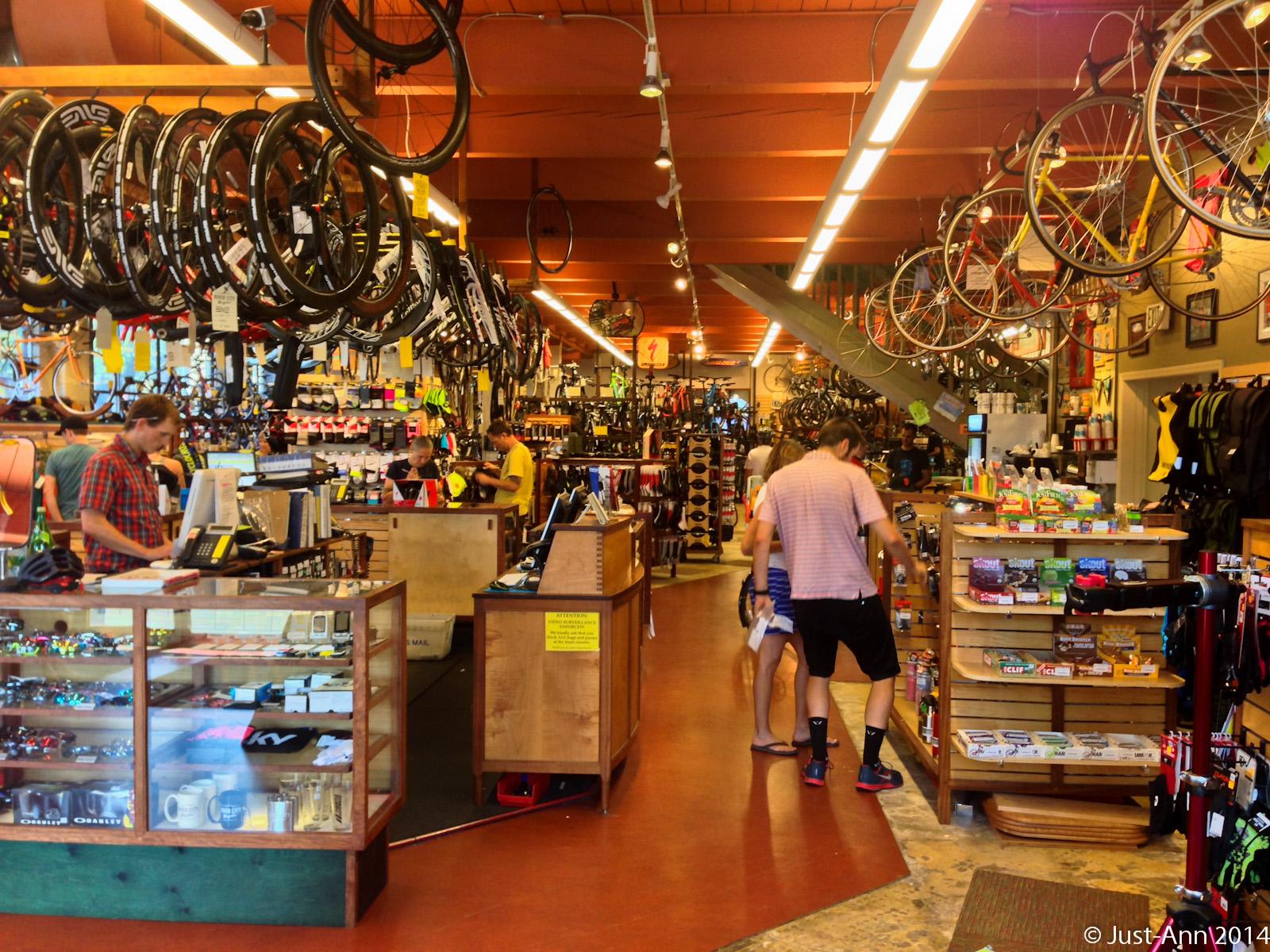 A busy bicycle shop interior filled with various bikes hanging from the ceiling, shelves stocked with cycling accessories, and customers browsing the merchandise. Two cash registers are visible, with a staff member assisting a customer. The environment is bright and colorful, featuring a variety of products related to cycling.