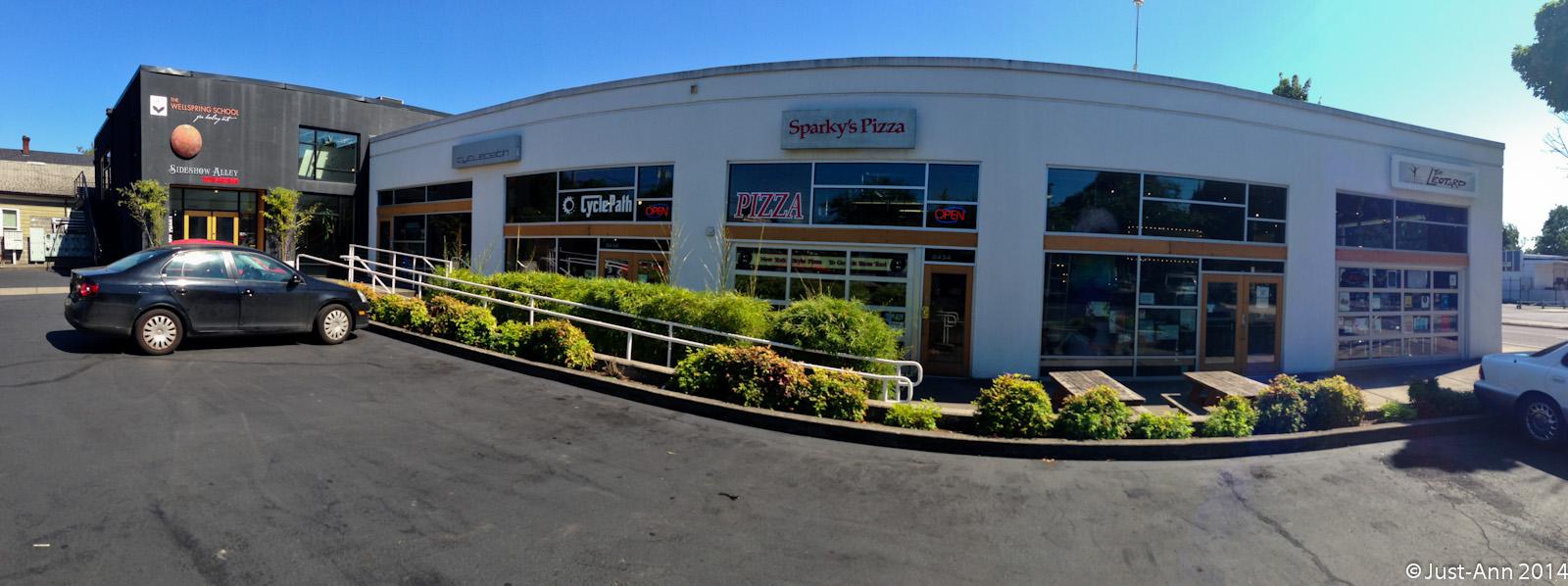 A panoramic view of a commercial area featuring Sparky's Pizza with a bright 'OPEN' sign. The building has large windows and is surrounded by well-maintained greenery. Nearby, the Sideshow Alley is visible on the left, alongside The Wellspring School building. A black car is parked in the foreground, with additional outdoor seating visible in front of the shops on a sunny day.