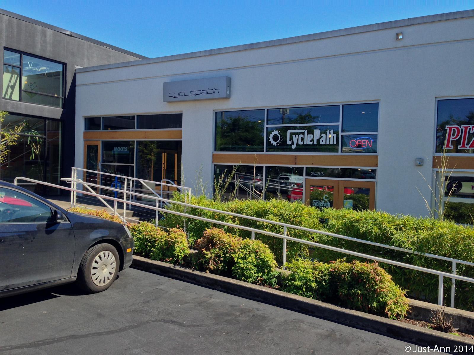 A storefront of a bike shop named "CyclePath," featuring large glass windows displaying the shop's interior. The sign above the entrance reads "cyclepath," and there is an illuminated "OPEN" sign in the window. In the foreground, a black car is parked, with a landscaped area featuring green plants and a railing leading up to the entrance. The sky is clear and blue, indicating a sunny day.