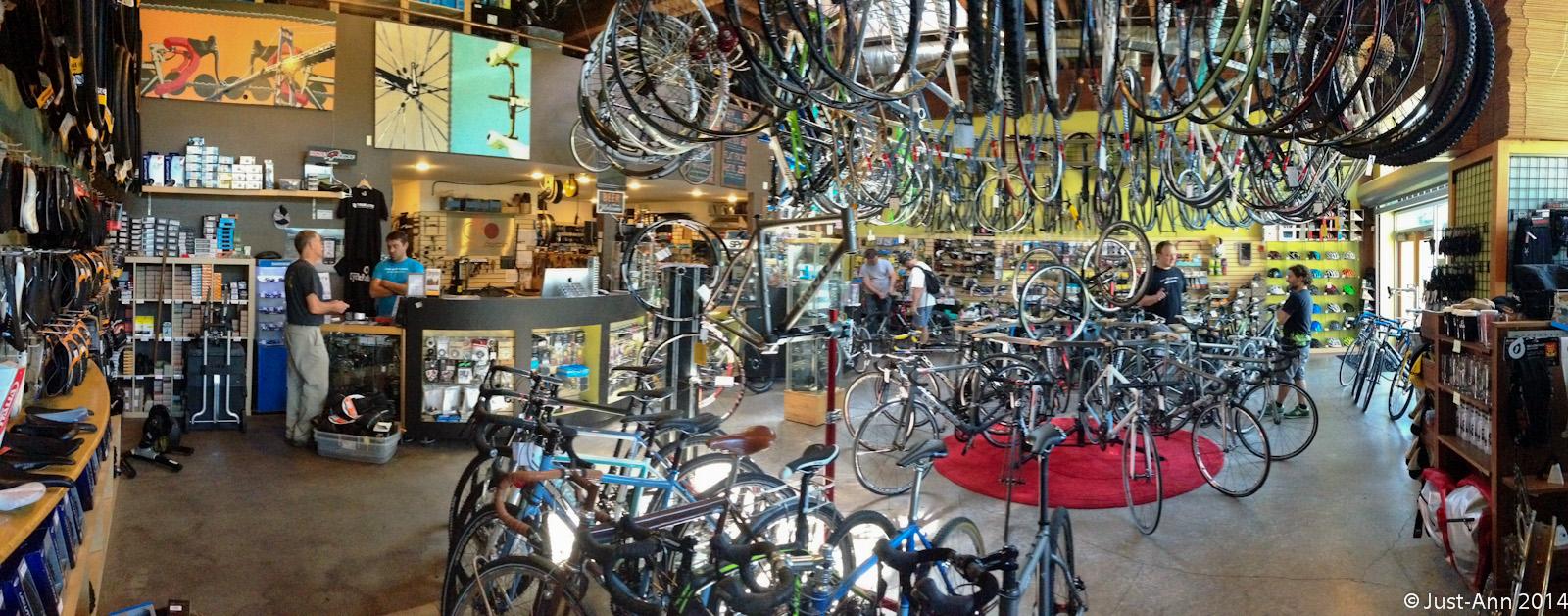 A panoramic view of a busy bike shop featuring various bicycles on display, with racks of bike parts and accessories lining the walls. Two staff members are engaged with customers at the checkout counter, while other shoppers explore the wide selection of bikes and gear. The shop has a warm, inviting atmosphere with colorful decorations and ample natural light.