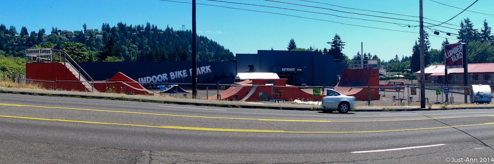 A panoramic view of an outdoor bike park featuring various ramps and half-pipes. The facility is located next to a busy road, with a large sign reading "Indoor Bike Park" visible on the building. Surrounding the park are trees and hills in the background, while additional signage indicates summer camps and other activities nearby. A few parked cars can be seen in the foreground. The day is sunny with clear blue skies. Lumberyard Indoor Bike Park mountain bike trail.