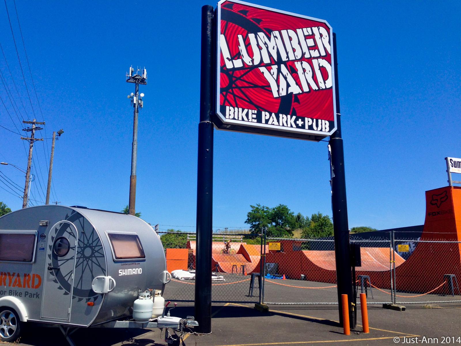 A sunny day at Lumber Yard Bike Park and Pub featuring a large, colorful sign displaying the park's name. In the foreground, a silver camper is parked beside the entrance, with a bicycle wheel graphic on its side. The bike park area includes various ramps and structures visible in the background, enclosed by a fence. Power lines and a clear blue sky complete the setting. Lumberyard Indoor Bike Park mountain bike trail.