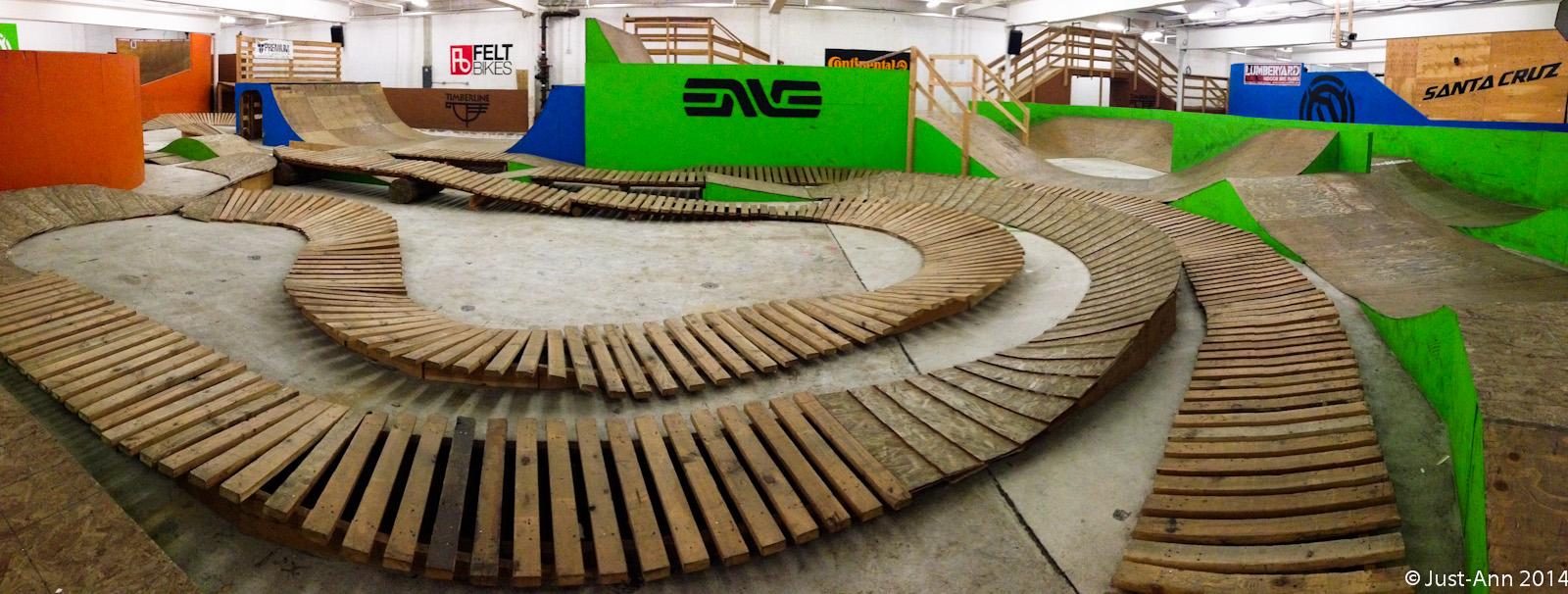 A panoramic view of an indoor bike park featuring various wooden tracks and ramps. The layout includes winding paths made of wooden planks and multiple sloped ramps painted in bright colors, including green and orange. The walls are adorned with logos from different bike brands, and the space is well-lit, providing an inviting atmosphere for biking enthusiasts. Lumberyard Indoor Bike Park mountain bike trail.