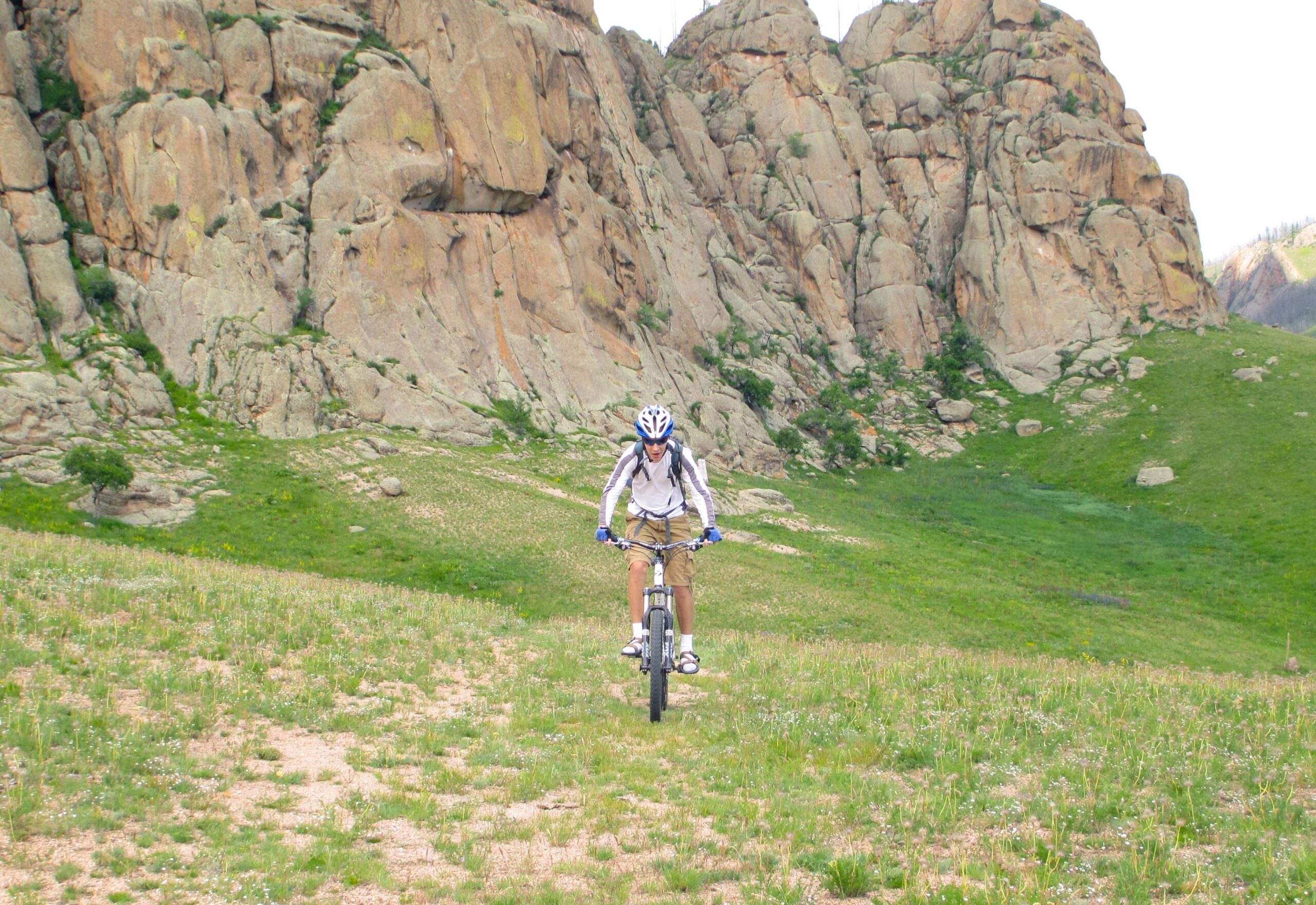 A person riding a mountain bike on a grassy path with rocky cliffs in the background, surrounded by green hills. Bearded Vulture Pass Trails mountain bike trail.
