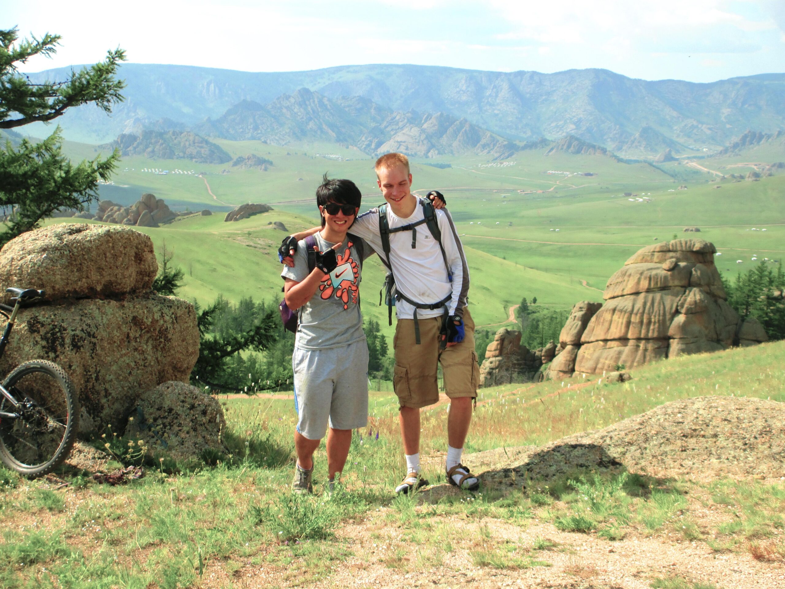 Two people standing together on a grassy hillside with a mountainous landscape in the background. One person is wearing a gray t-shirt and shorts, while the other is dressed in a long-sleeve shirt and shorts. They are smiling and posing for the photo, with a bicycle visible nearby and large rock formations in the distance. Bearded Vulture Pass Trails mountain bike trail.