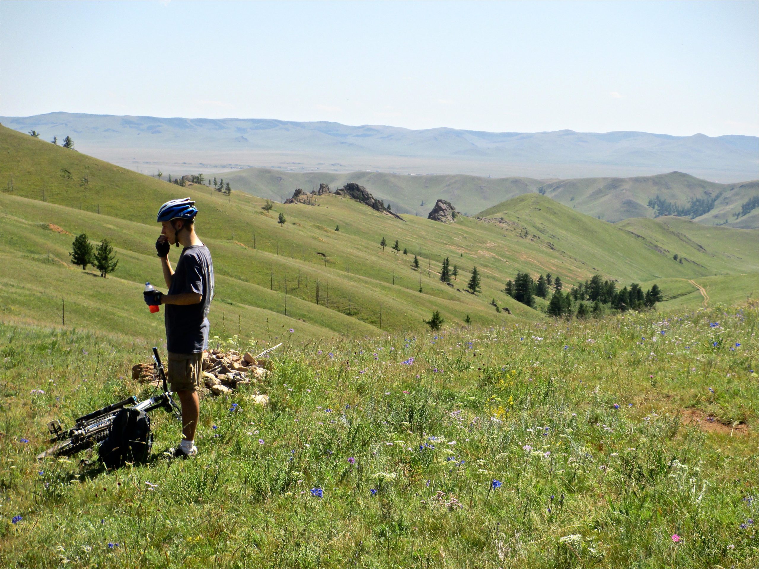 A cyclist in a helmet stands on a grassy hillside, enjoying a drink while taking in the scenic view of rolling green hills and distant mountains under a clear blue sky. A mountain bike is nearby, surrounded by wildflowers. Camel Run mountain bike trail.