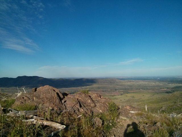 A panoramic view of a mountainous landscape with rocky foreground and a wide-open sky. The scene captures rolling hills and valleys stretching into the distance, with scattered vegetation and remnants of fallen branches visible. The sky is clear with soft clouds, creating a serene atmosphere. Ginny Trail mountain bike trail.