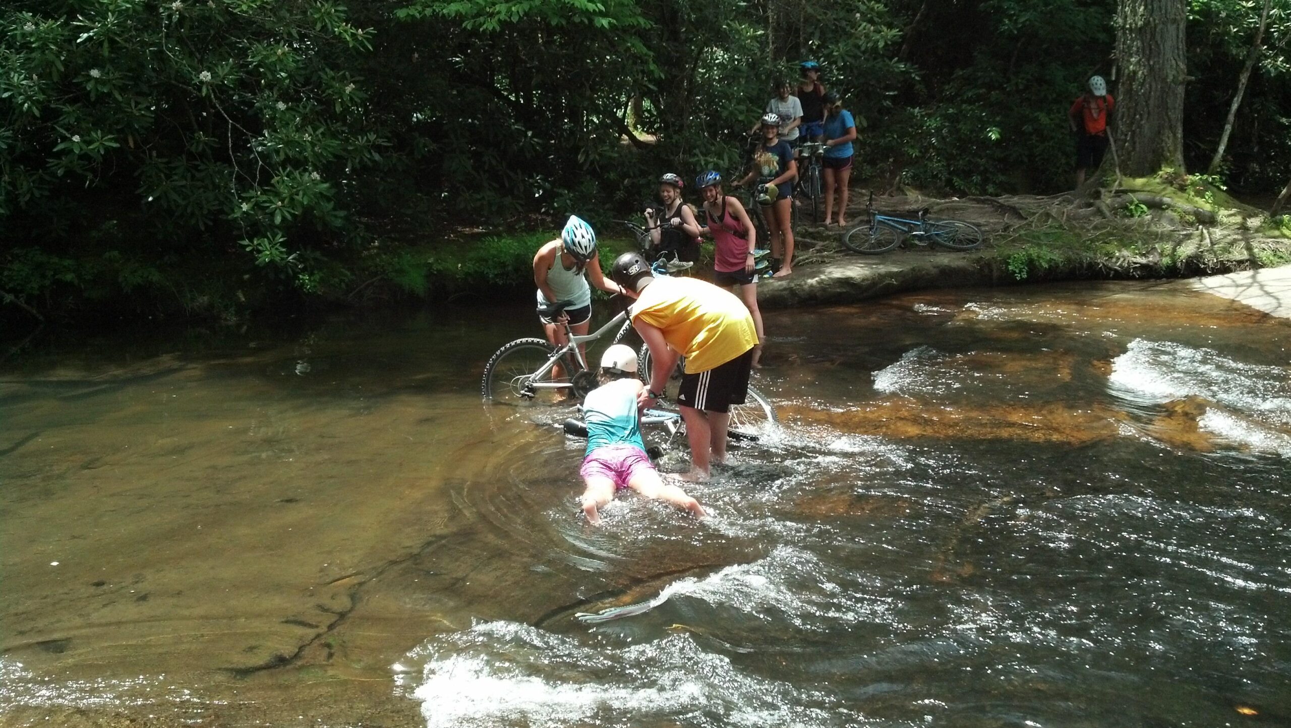 A group of people is gathered by a shallow stream, with several bicycles nearby. Two individuals are attempting to lift a bicycle out of the water while a child sits in the stream, partially submerged. Other onlookers, including a few wearing helmets, are watching the scene from the bank. Lush greenery surrounds the area, creating a vibrant outdoor setting. DuPont State Forest mountain bike trail.