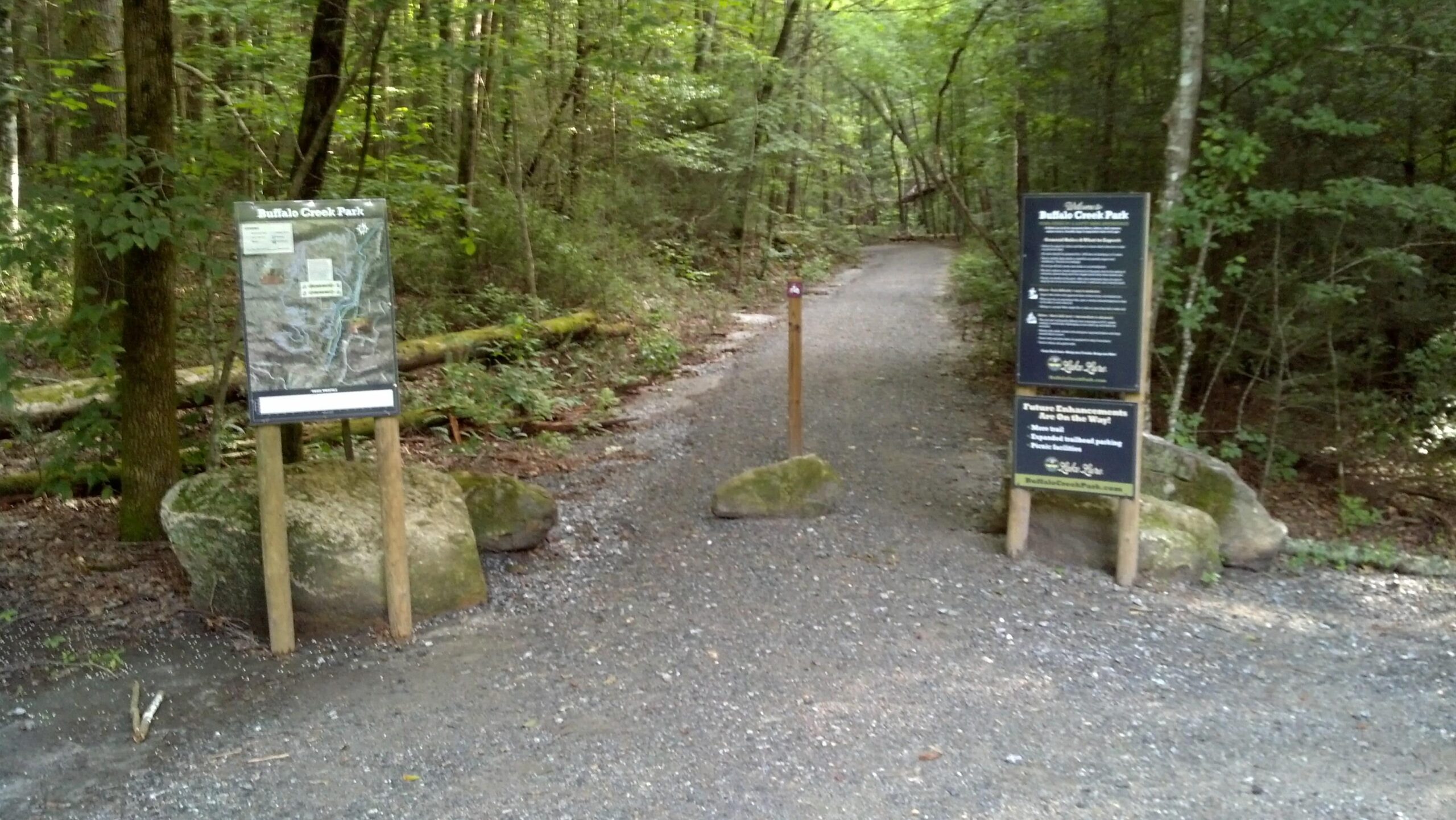 A gravel pathway leading into a wooded area, marked by two informational signs at the entrance of Buffalo Creek Park. The signs provide a map and park guidelines, flanked by large stones on either side of the trail. Lush green trees and foliage surround the path, creating a serene natural environment. Buffalo Creek Park: The Head, The Heart &amp; The Tail (HHT) mountain bike trail.