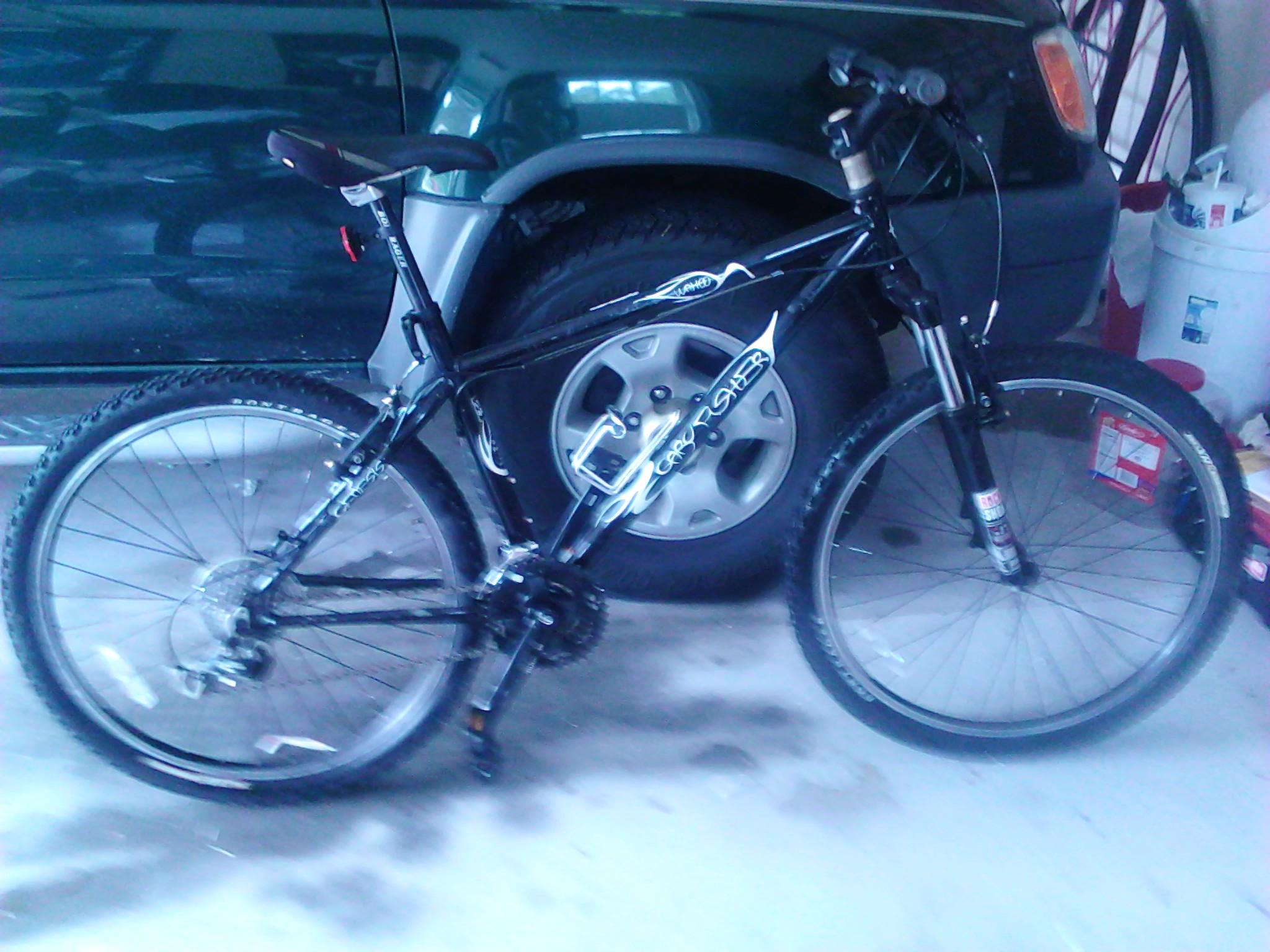 Gary Fisher Wahoo: A black mountain bike is positioned in a garage next to a green SUV, with tools and other items visible in the background. The bike features thick tires, a gear system, and a standard bike seat. The garage floor is concrete and slightly debris-ridden.