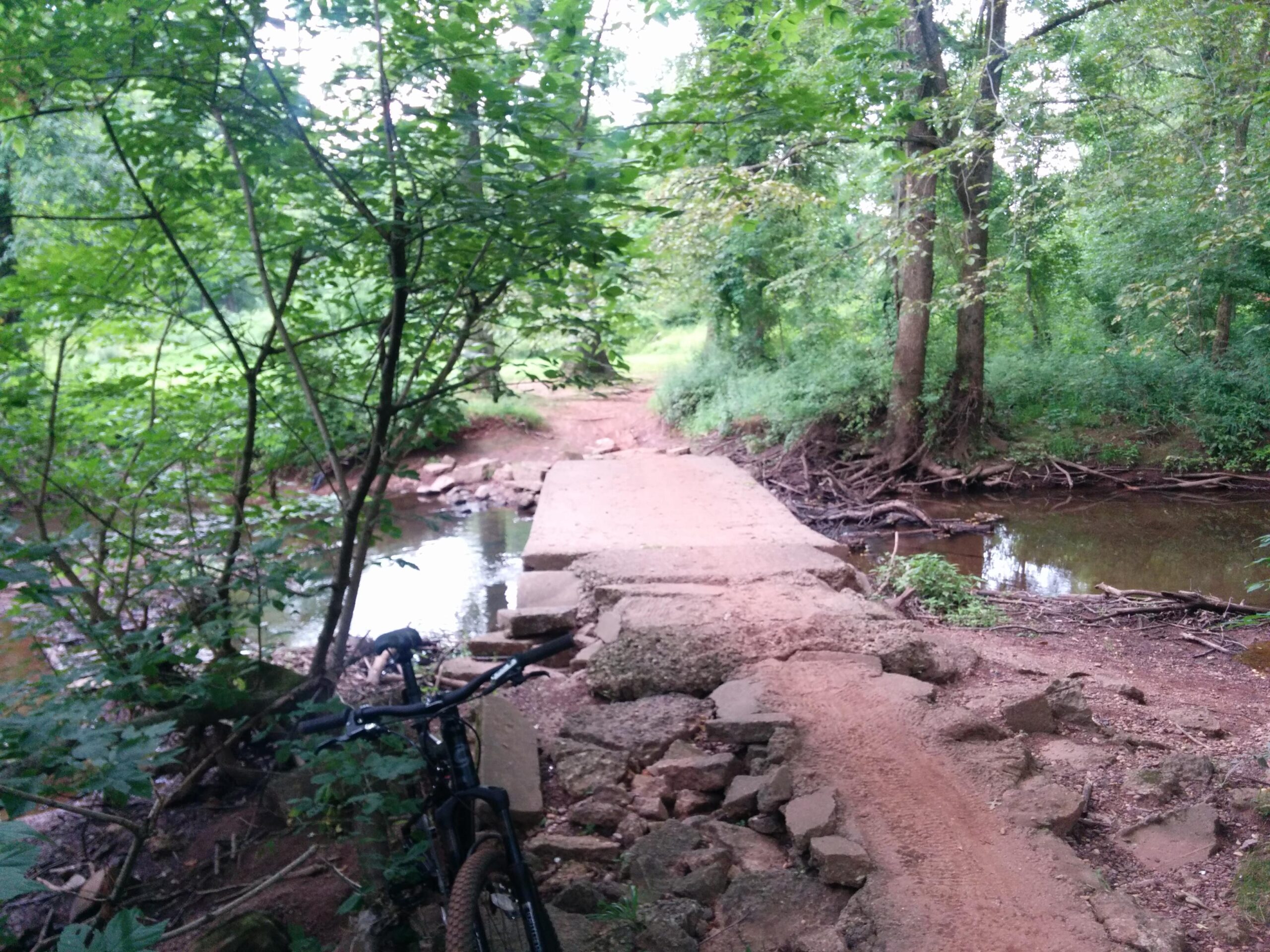 A rocky bridge over a small creek surrounded by lush greenery, with a mountain bike leaning against the left side. The path leads into a wooded area in the background. Six Mile Run mountain bike trail.