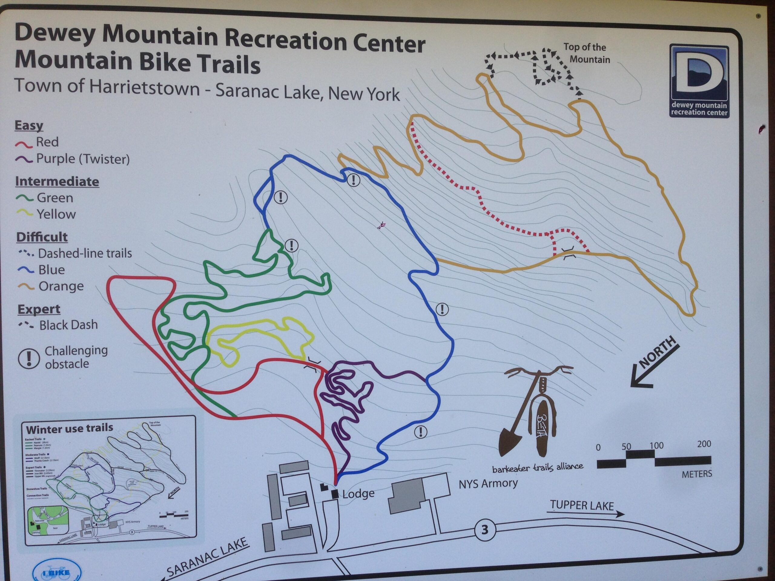 Map of Dewey Mountain Recreation Center's mountain bike trails in Saranac Lake, New York. It shows various trail difficulties color-coded: easy (red, purple), intermediate (green, yellow), difficult (blue, orange, dashed lines), and expert (black dashed). Includes a winter trail map inset and indicators for features like obstacles and the lodge location. Additionally, it marks "Top of the Mountain" and directional north. Dewey Mountain mountain bike trail.