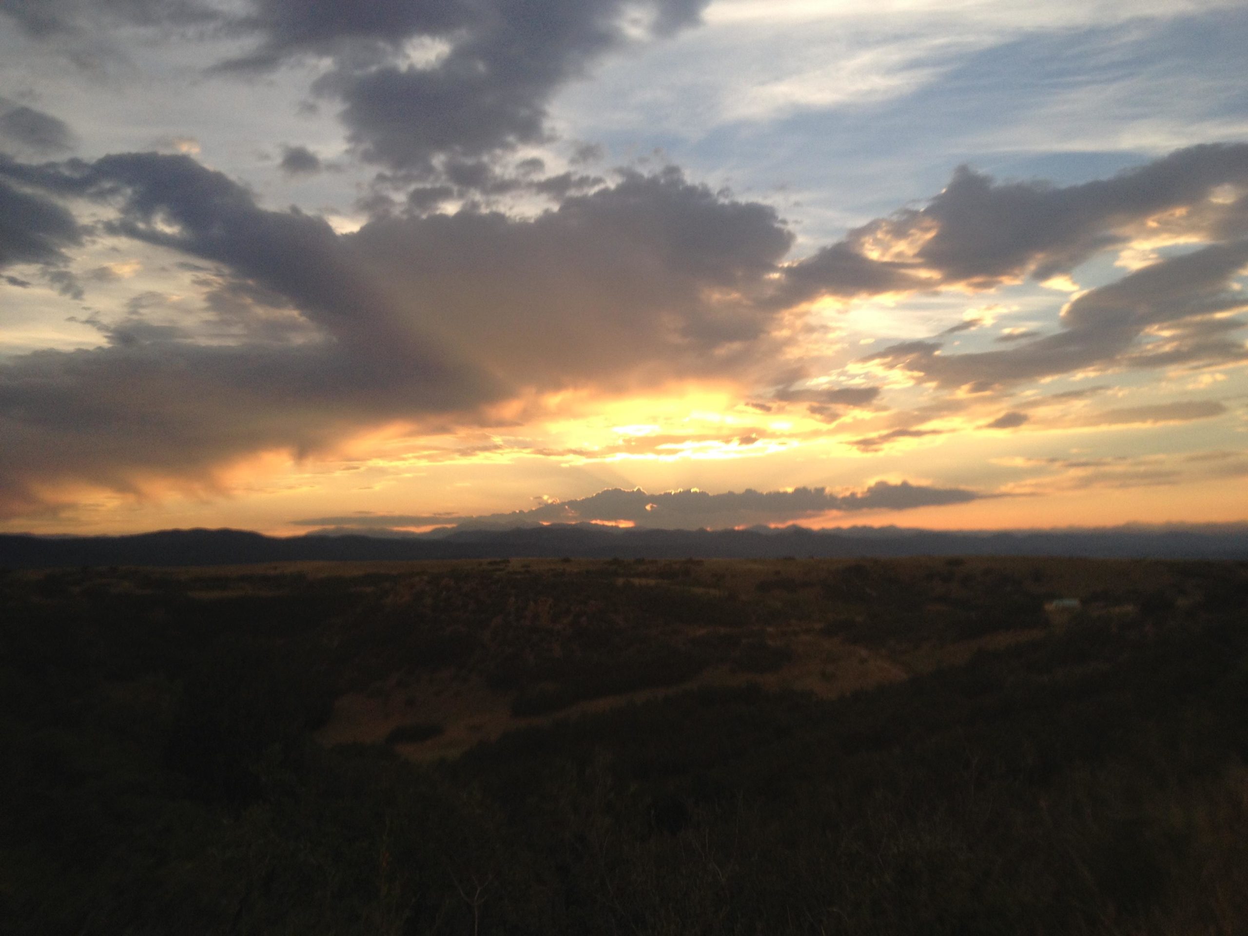 A scenic view of a sunset, featuring a colorful sky with clouds in shades of orange, pink, and gray. The sun is setting behind distant mountains, casting dramatic shadows across the landscape. The foreground shows rolling hills and patches of greenery beneath the expansive sky. Ridgeline Open Space Trail mountain bike trail.
