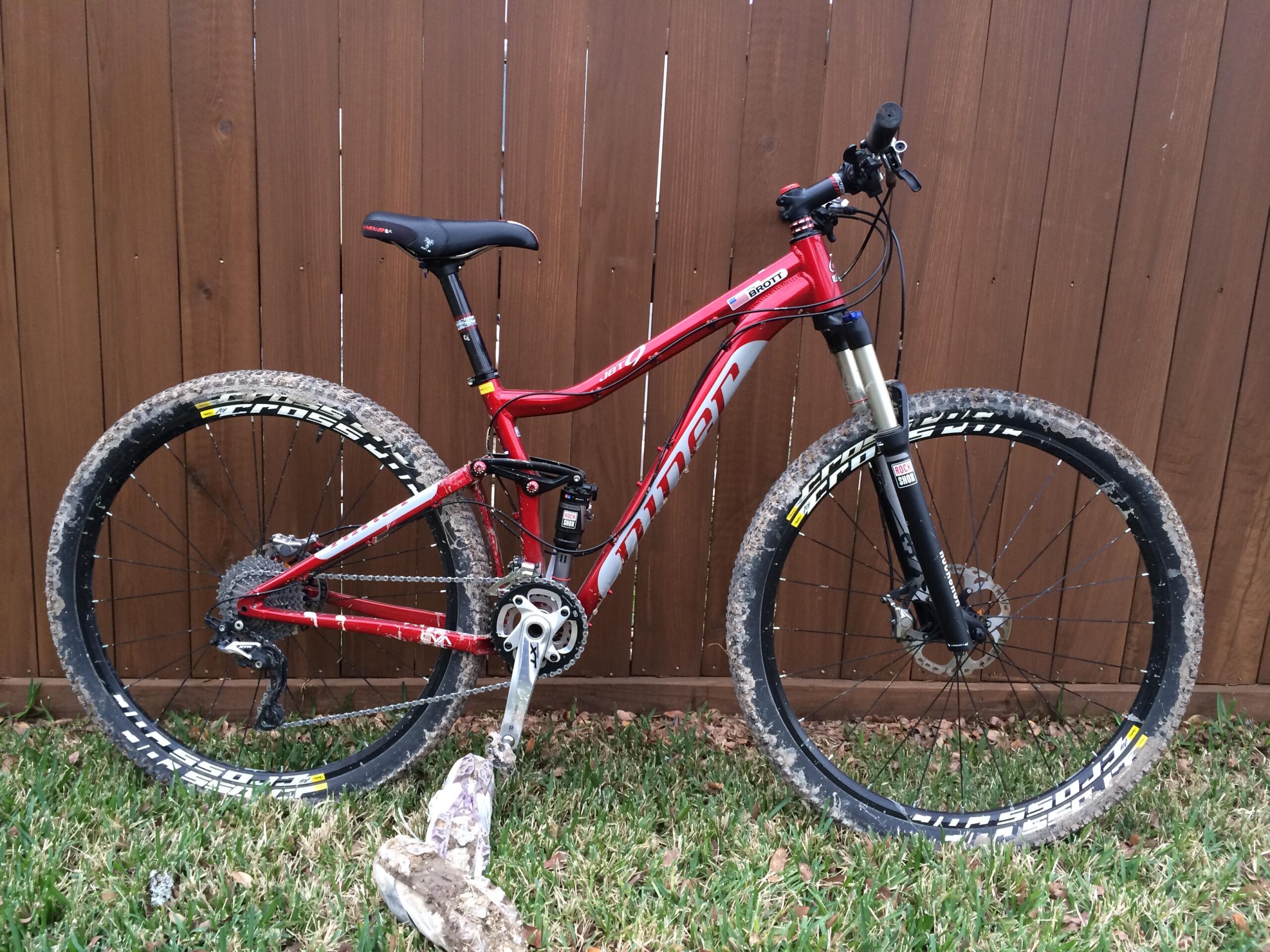 Niner Jet 9: A red mountain bike leaning against a wooden fence, featuring muddy tires and a sleek design. The bike has a prominent suspension fork, a black seat, and is positioned on green grass with scattered leaves.