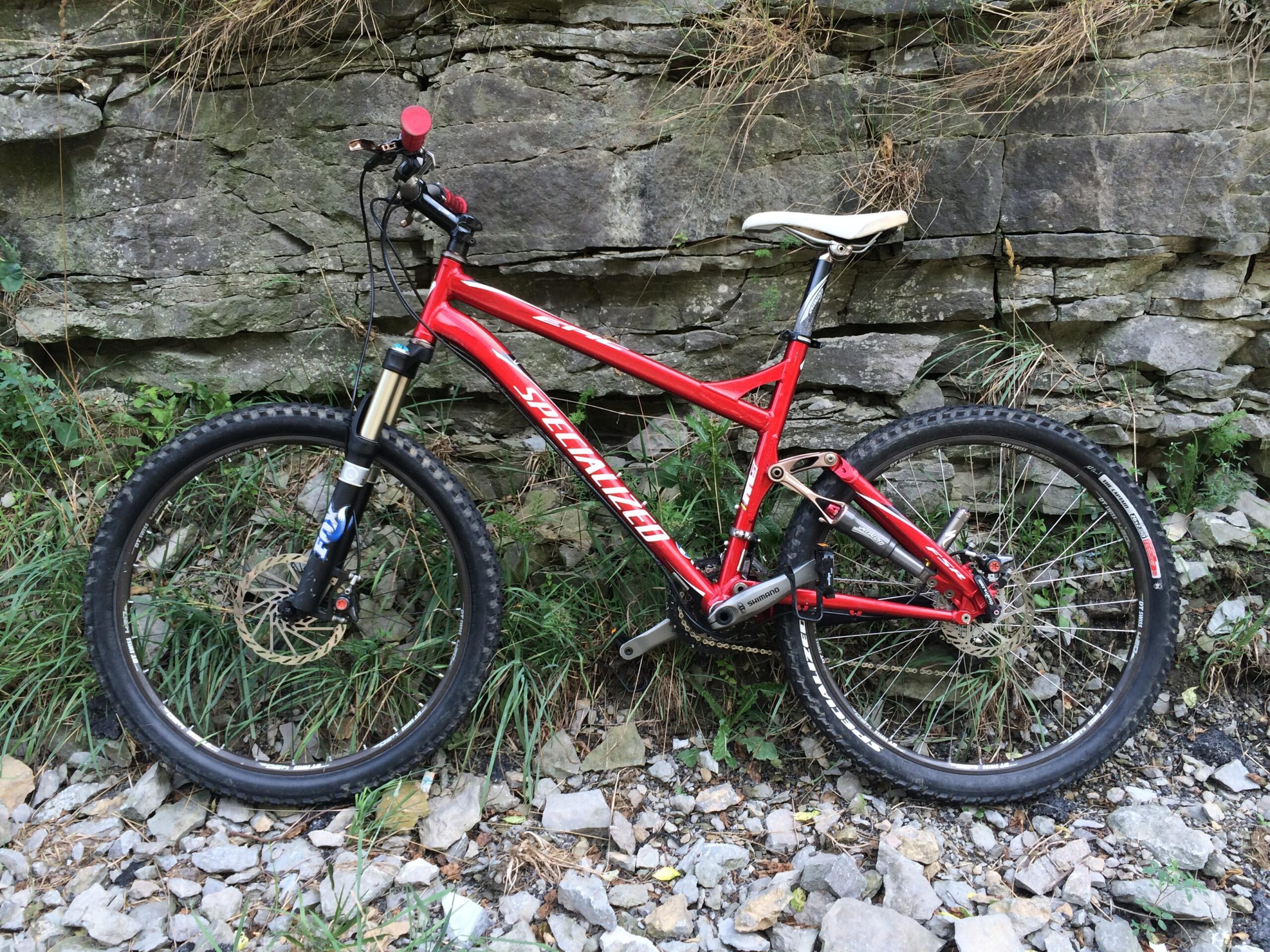 Specialized Epic Expert: A red mountain bike leaning against a rocky wall, surrounded by grass and small stones. The bike features thick tires, a suspension fork, and a contrasting white saddle.