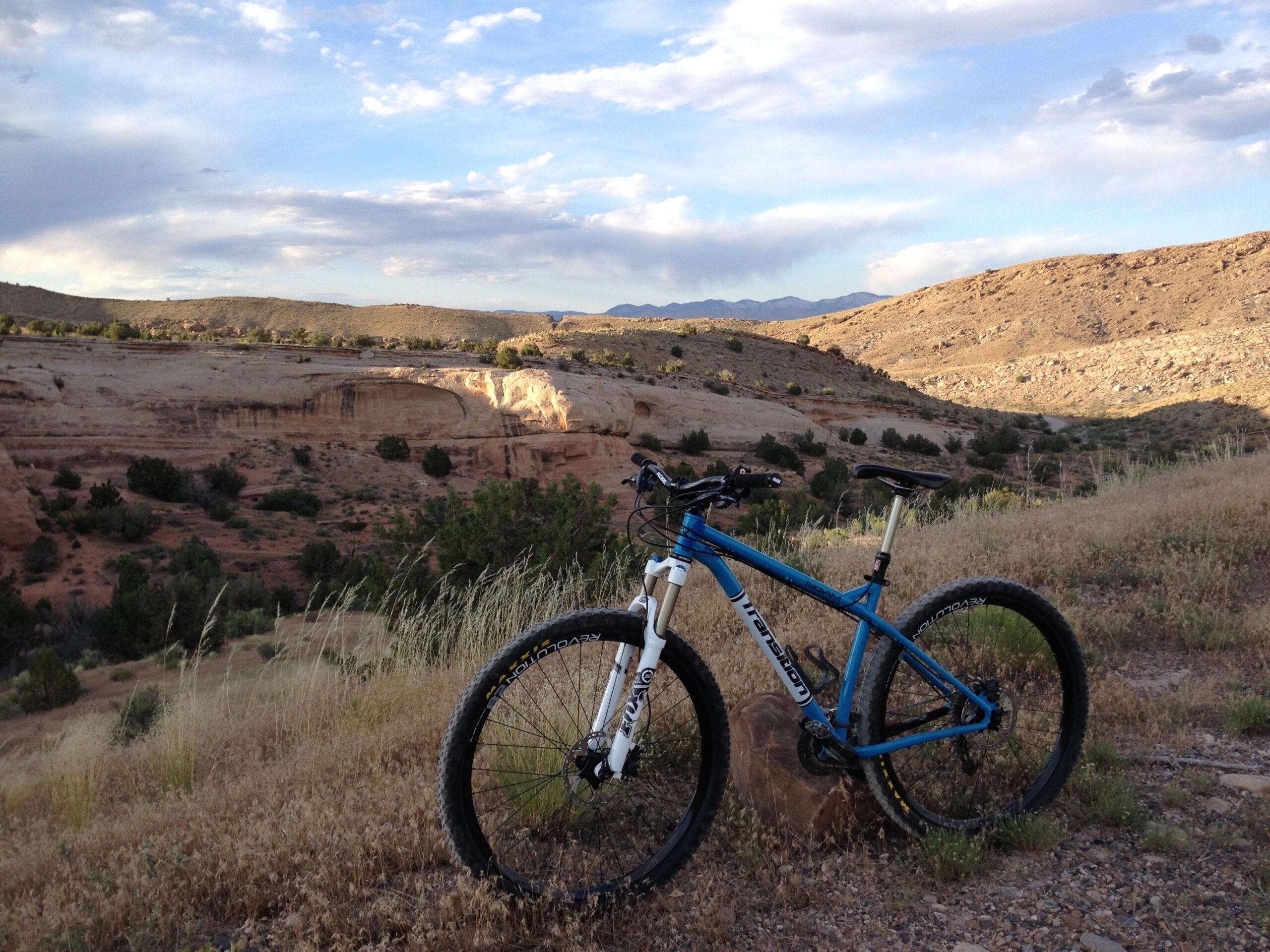 Transition TransAM: A blue mountain bike resting on a rock in a grassy area, with rolling hills and rocky cliffs in the background under a partly cloudy sky.