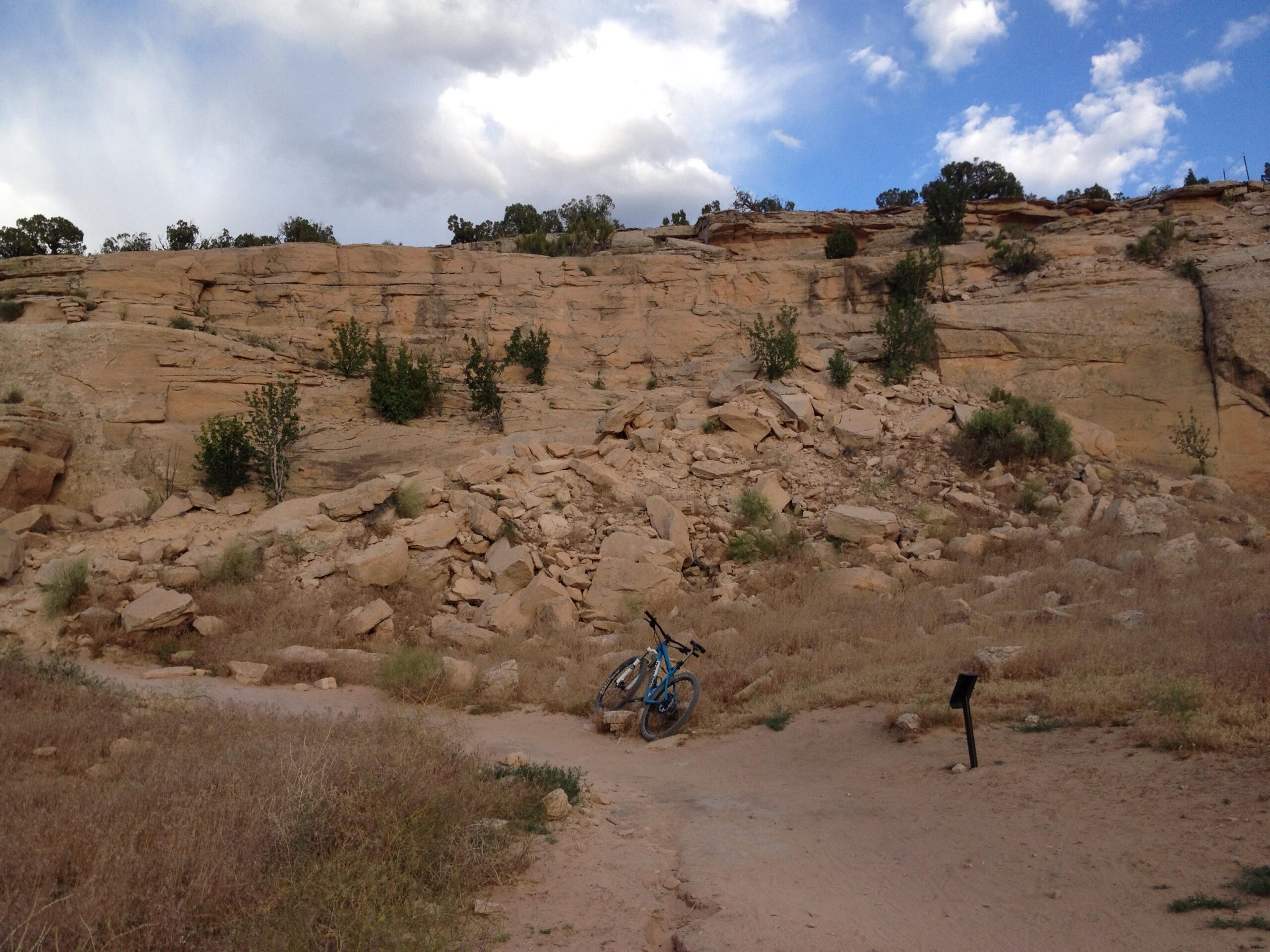 A rocky terrain featuring a steep cliff face with scattered vegetation, including small trees and shrubs. In the foreground, a blue mountain bike is leaned against a large rock, with a dirt path leading through dry grass and rocky ground. The sky is partly cloudy, indicating a mix of sunlight and clouds. Mary's Loop / Horsethief Bench mountain bike trail.