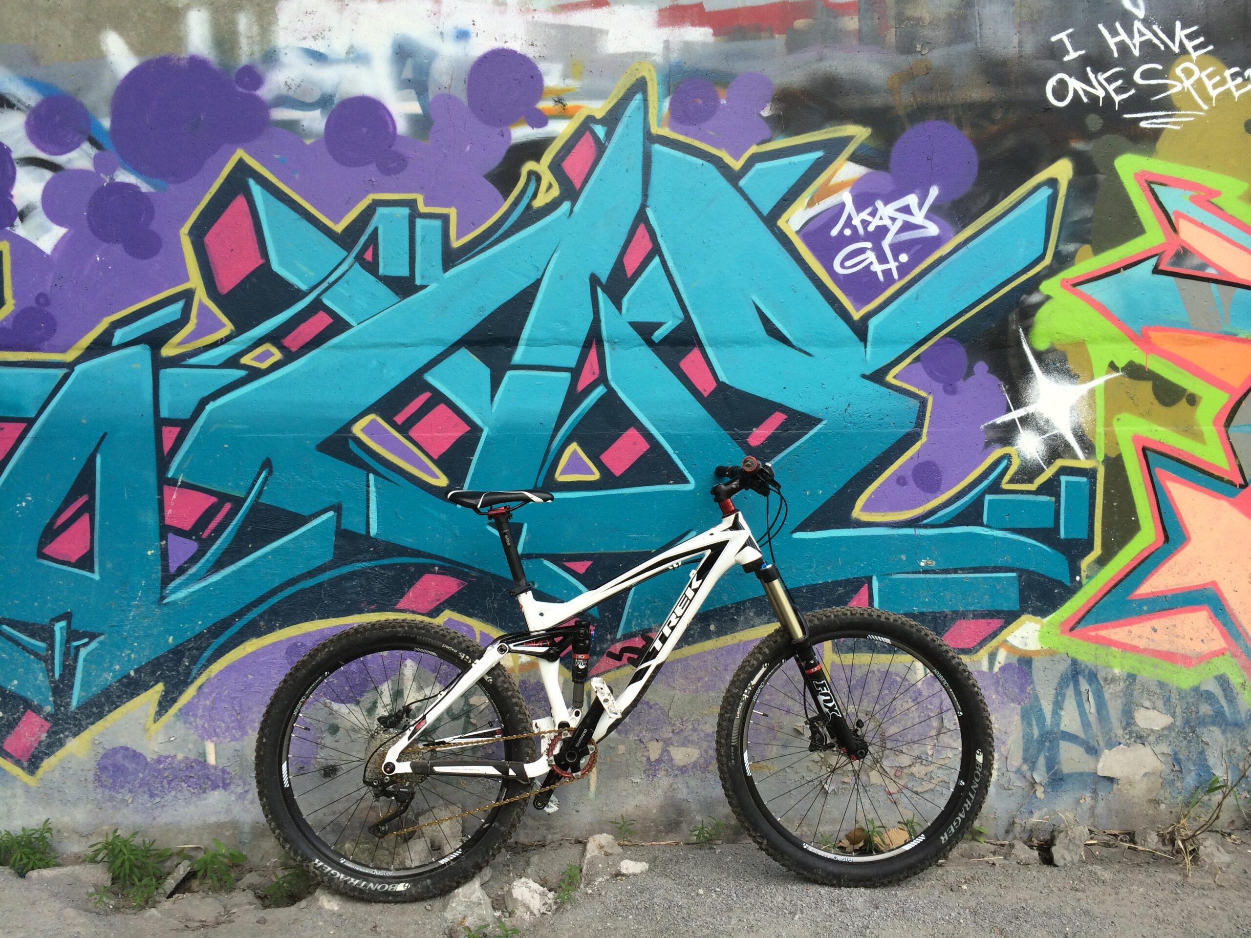 Trek Remedy 8: A white mountain bike leaning against a colorful graffiti wall featuring vibrant blue, purple, and pink abstract designs, with text and artistic elements. The bike's wheel and frame are clearly visible, showcasing its design against the urban backdrop.